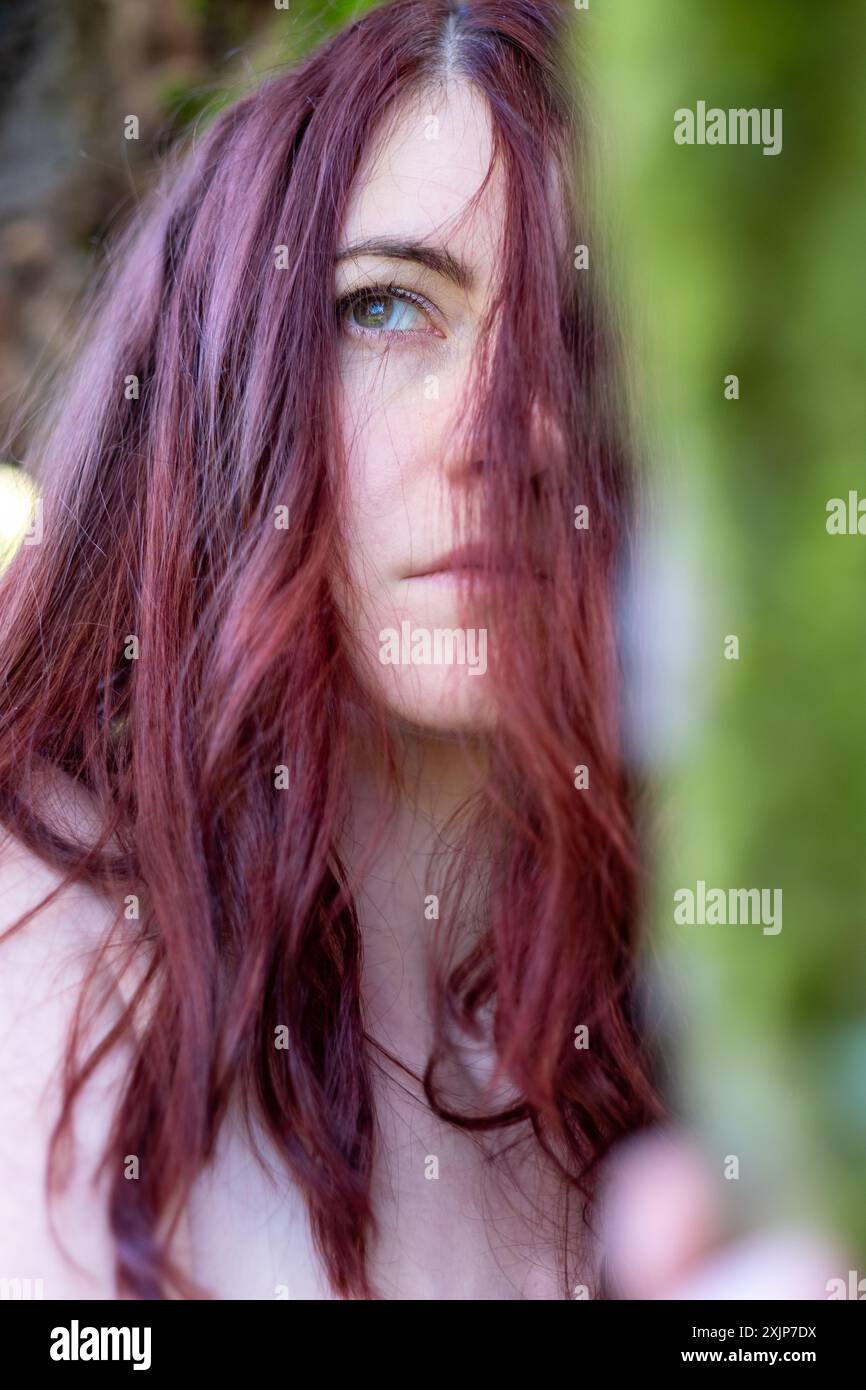 Una donna con lunghi capelli rossi copre parzialmente il viso, i suoi occhi blu che sbirciano attraverso i fili. Il primo piano cattura il suo intenso e contemplativo espr Foto Stock