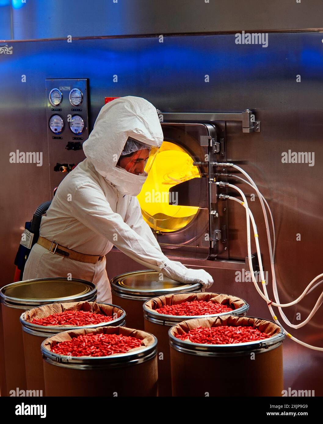 Un operatore che indossa guanti e indumenti protettivi maneggia fusti riempiti di capsule rosse brillanti all'interno di un impianto di produzione farmaceutica. Il Wo Foto Stock