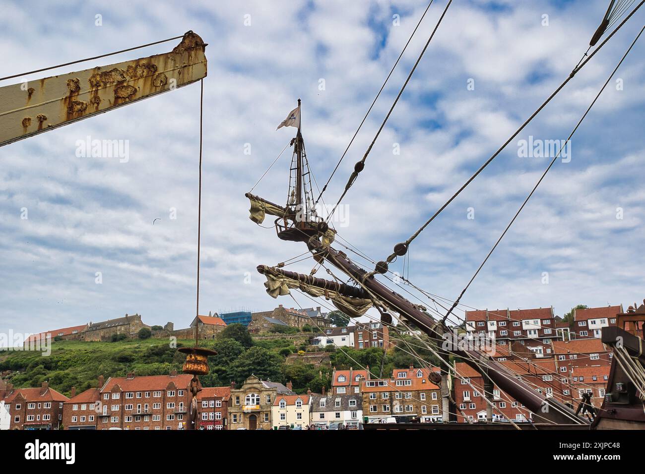 Vista dell'albero e dell'attrezzatura di una nave storica con una città costiera e una collina sullo sfondo. Il cielo è parzialmente nuvoloso. Foto Stock
