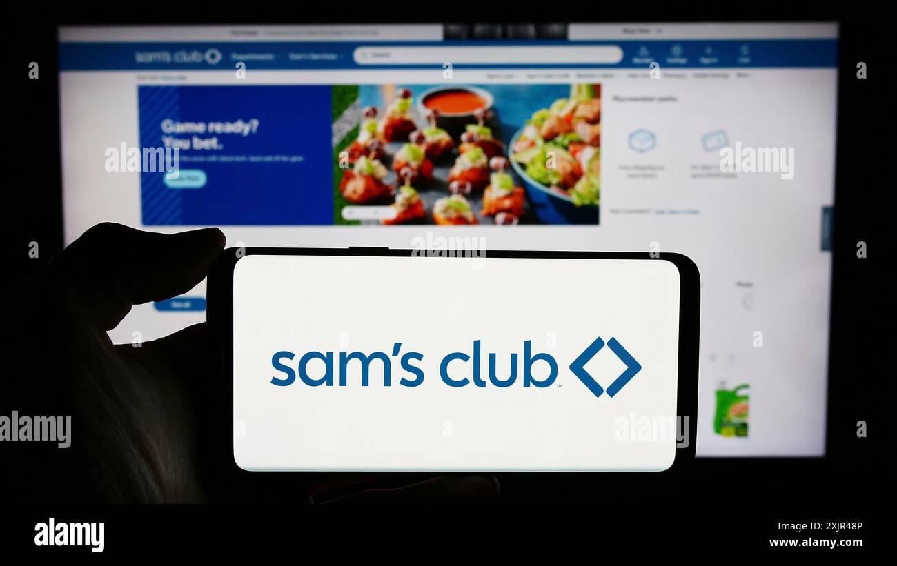 Stoccarda, Germania, 02-07-2023: Persona che detiene un cellulare con il logo della società di negozi al dettaglio Sam's Club, solo affiliata agli Stati Uniti, davanti all'azienda Foto Stock