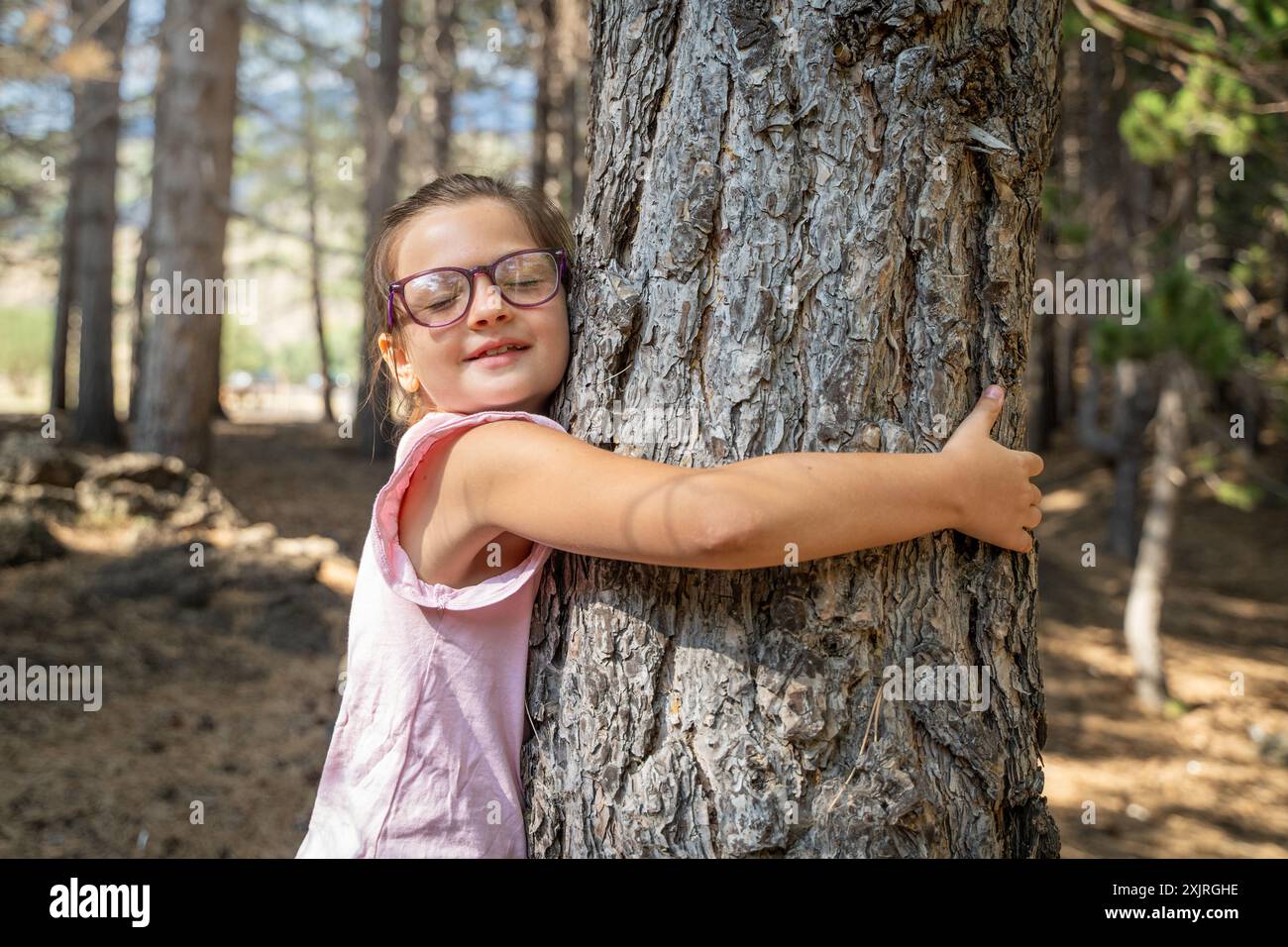 Giovane ragazza con occhiali che abbraccia un grande tronco di albero in una foresta illuminata dal sole. Bambino che mostra amore per la natura - consapevolezza ambientale e importanza del pre Foto Stock