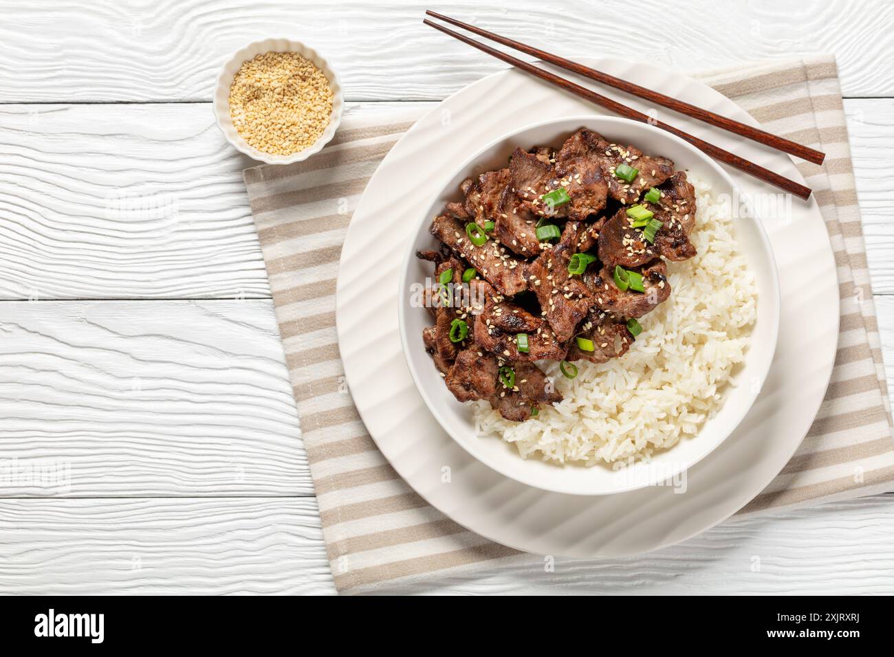 Bulgogi, manzo BBQ coreano, manzo fritto a fette sottili cosparso di semi di sesamo e cipolla di primavera servito con riso in ciotola su un tavolo di legno bianco con tritatutto Foto Stock