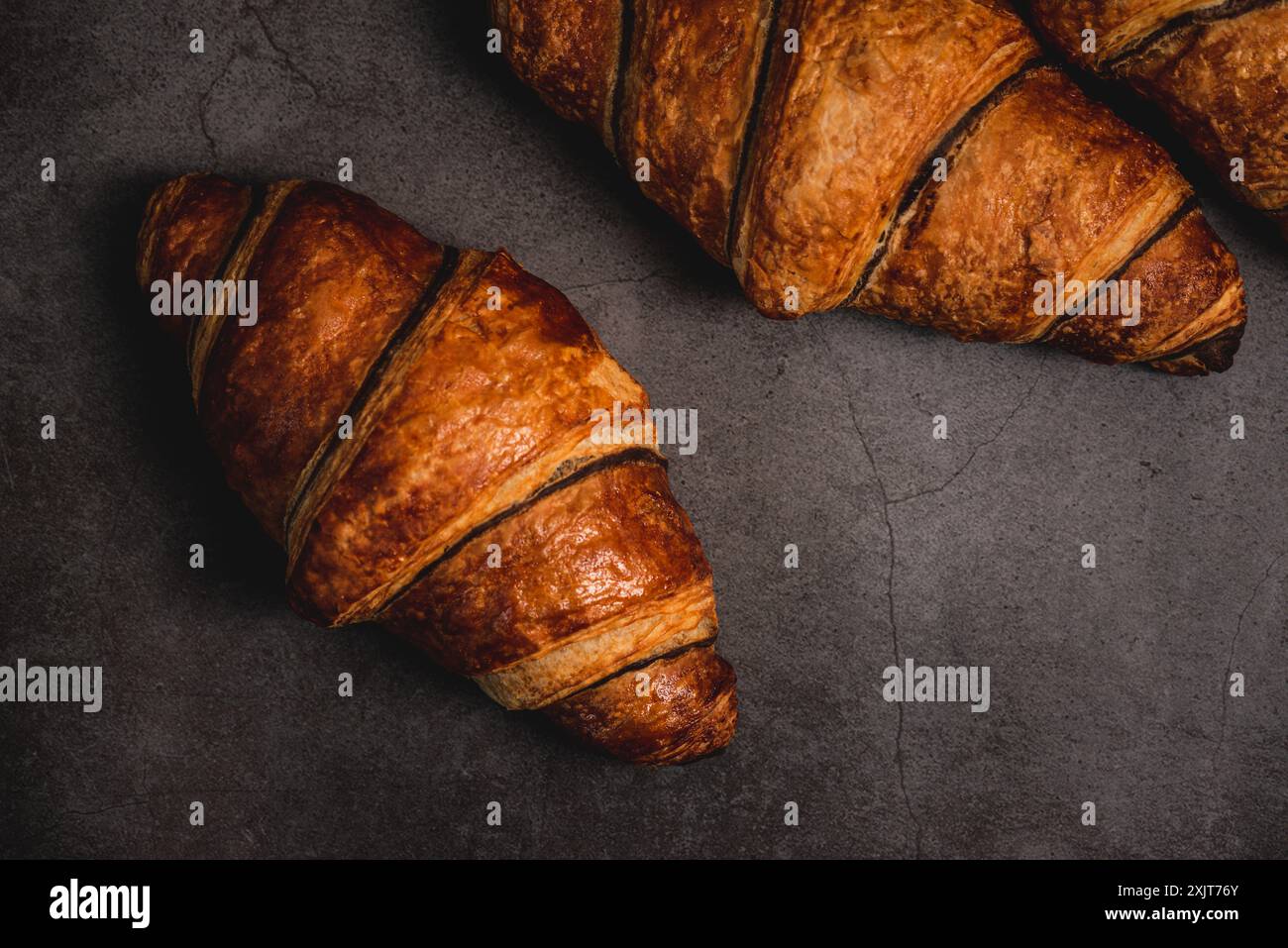 Tre croissant appena sfornati si trovano su un ripiano grigio scuro, pronti per essere divorati. La crosta marrone-dorata risplende sotto la luce soffusa, invitando a Foto Stock