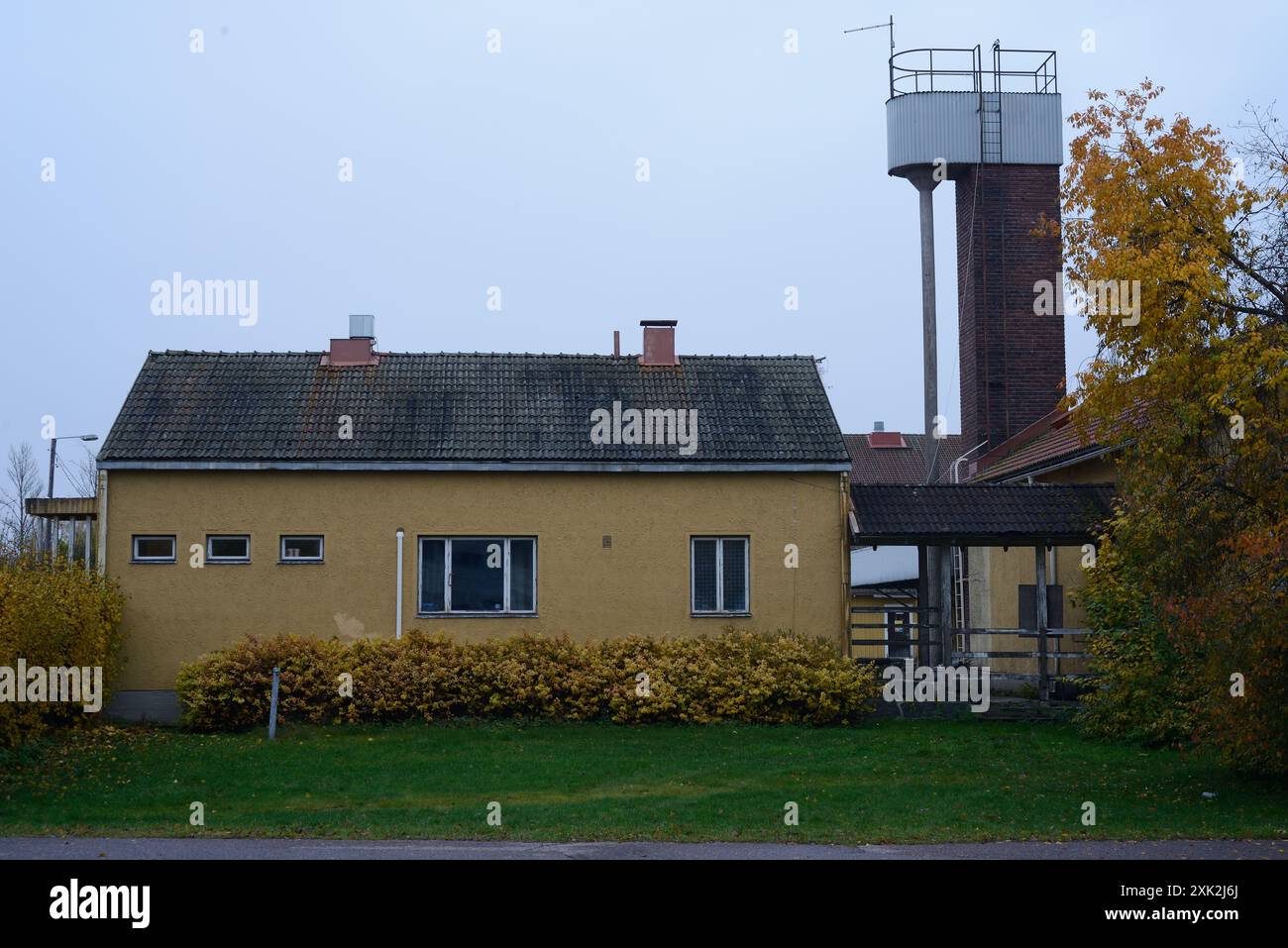 Un semplice edificio giallo a un piano con un tetto piastrellato e piccole finestre rettangolari, adagiate su un cielo coperto. L'edificio è fiancheggiato dall'autunno Foto Stock