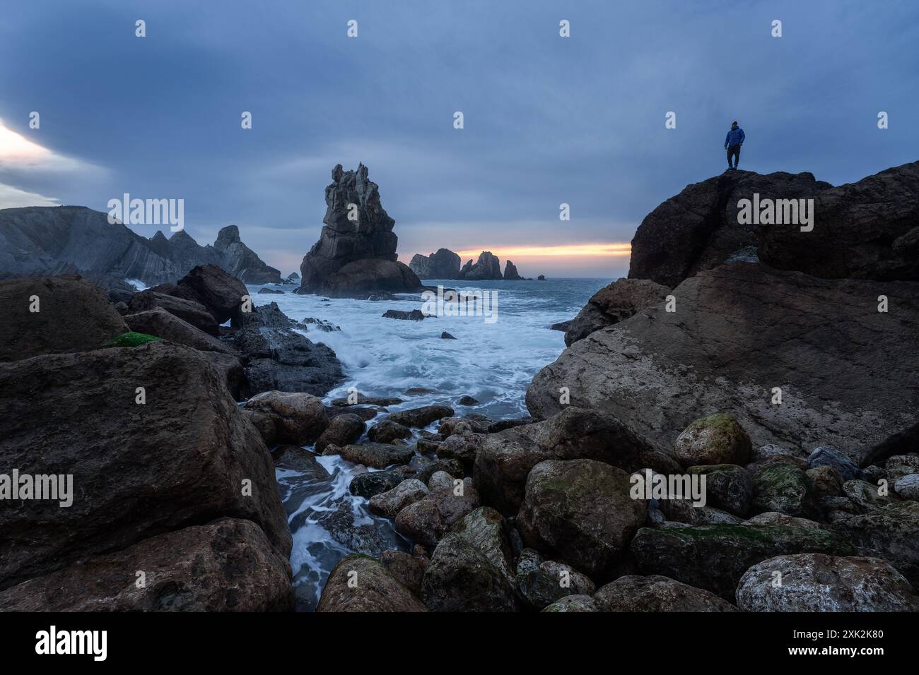 Un esploratore si erge sulla costa frastagliata e rocciosa della Costa Quebrada in Cantabria, Spagna, e si affaccia sulle spettacolari formazioni rocciose sotto un sottosuolo Foto Stock
