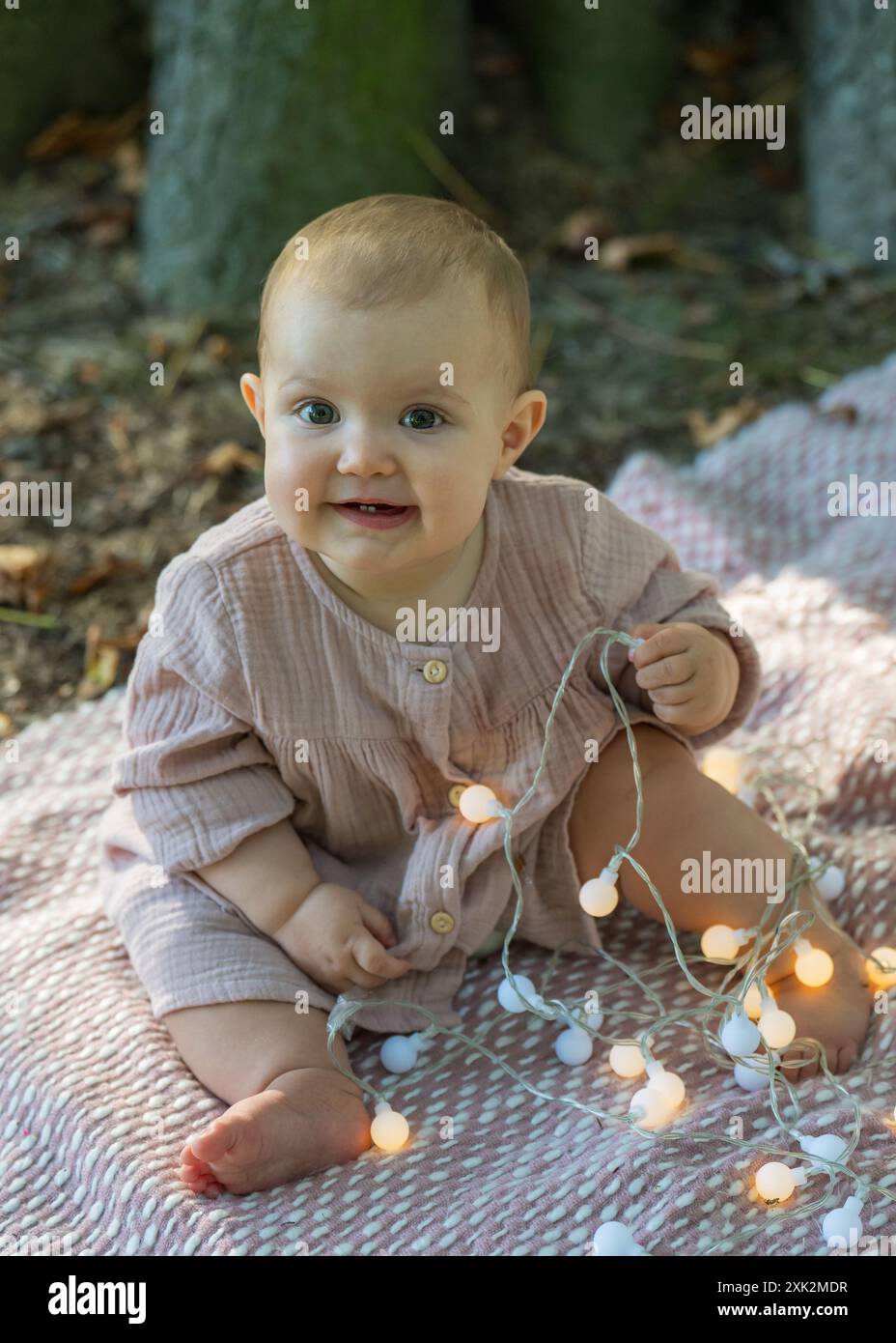 Ritratto di una bambina affascinante in un abito beige seduta vicino a un grande albero, guardando la macchina fotografica. Tiene le lanterne e le mastica con piacere. Foto Stock