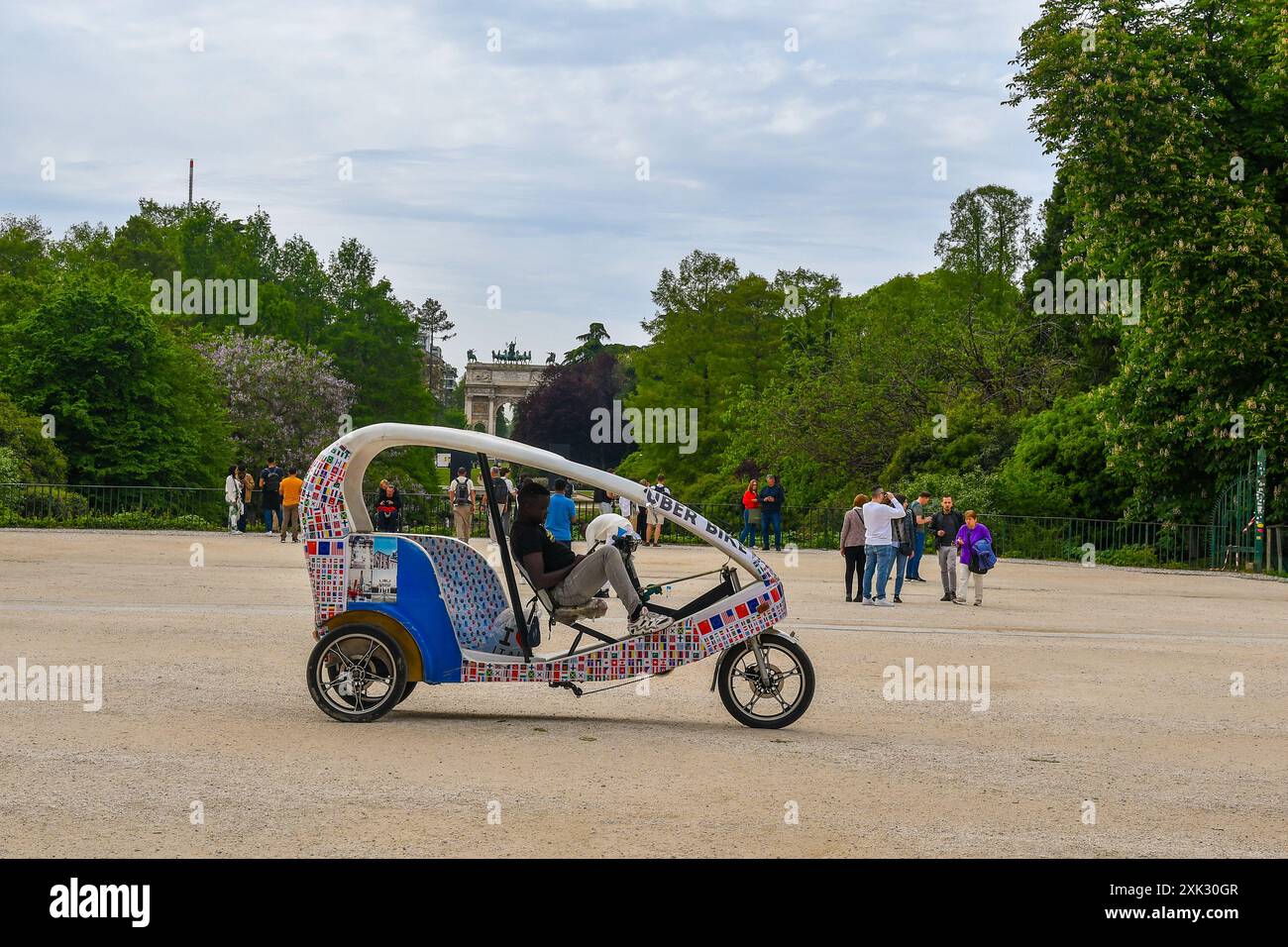 Un moderno risciò a tre ruote in attesa di passeggeri nel Parco Sempione con l'Arco della Pace sullo sfondo in primavera, Milano, Italia Foto Stock