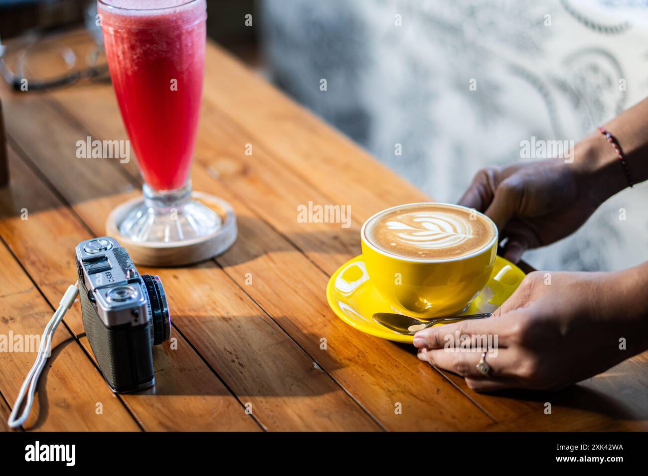 Una persona che posiziona una tazza gialla di latte art su un tavolo di legno accanto a una bevanda rosa in un bicchiere alto. Una fotocamera vintage è anche sul tavolo, creando Foto Stock