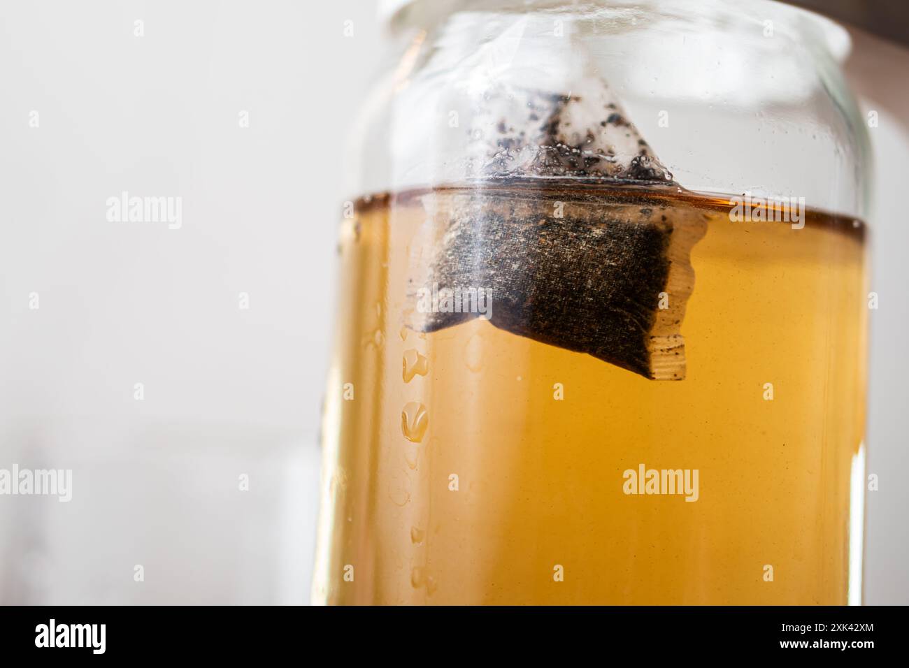 Primo piano di un vaso di vetro contenente il tè estratto, con un sacchetto di tè immerso nel liquido. Il tè è di colore ambra intenso e il vaso è parzialmente fille Foto Stock Primo piano di un vaso di vetro contenente il tè estratto, con un sacchetto di tè immerso nel liquido. Il tè è di colore ambra intenso e il vaso è parzialmente fille Foto Stock