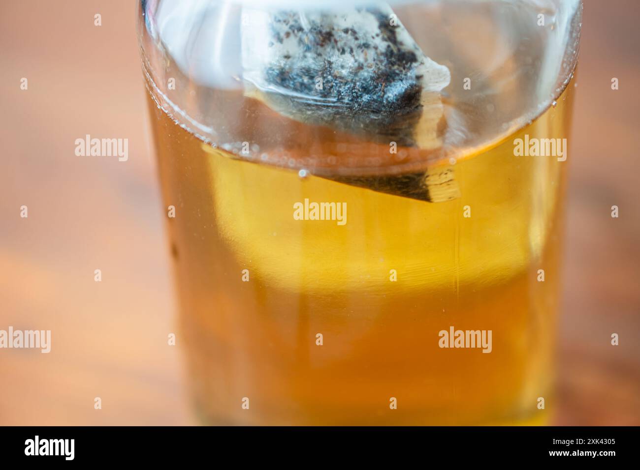 Un primo piano di un vaso di vetro contenente un liquido dorato, probabilmente tè, con una bustina di tè immersa all'interno. Il vaso è posizionato su una superficie di legno, in modo da mostrare Foto Stock Un primo piano di un vaso di vetro contenente un liquido dorato, probabilmente tè, con una bustina di tè immersa all'interno. Il vaso è posizionato su una superficie di legno, in modo da mostrare Foto Stock