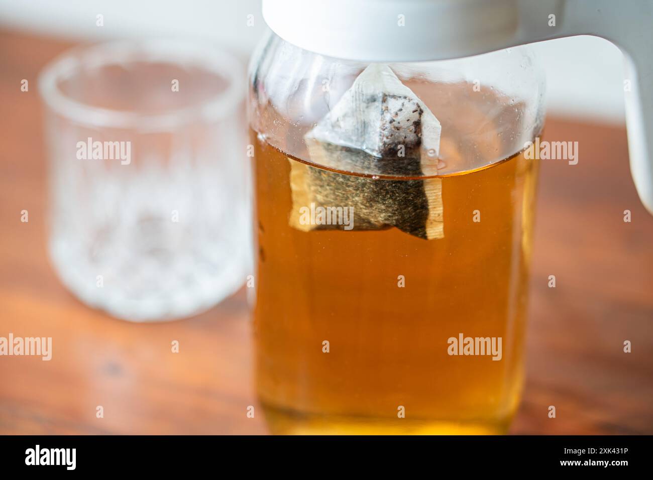 Un vaso di vetro riempito di tè estratto, con una bustina di tè all'interno. Il vaso è posizionato su una superficie di legno, con un bicchiere di vetro trasparente sul retro Foto Stock Un vaso di vetro riempito di tè estratto, con una bustina di tè all'interno. Il vaso è posizionato su una superficie di legno, con un bicchiere di vetro trasparente sul retro Foto Stock