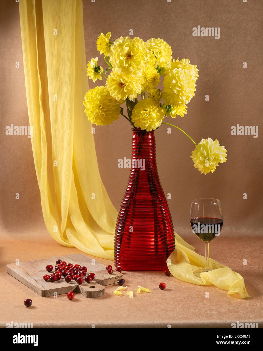 Un grande bouquet di fiori gialli in un vaso rosso, accanto a un bicchiere di vino rosso e frutti di bosco rossi su sfondo arancione e a un pendente scialle giallo Foto Stock