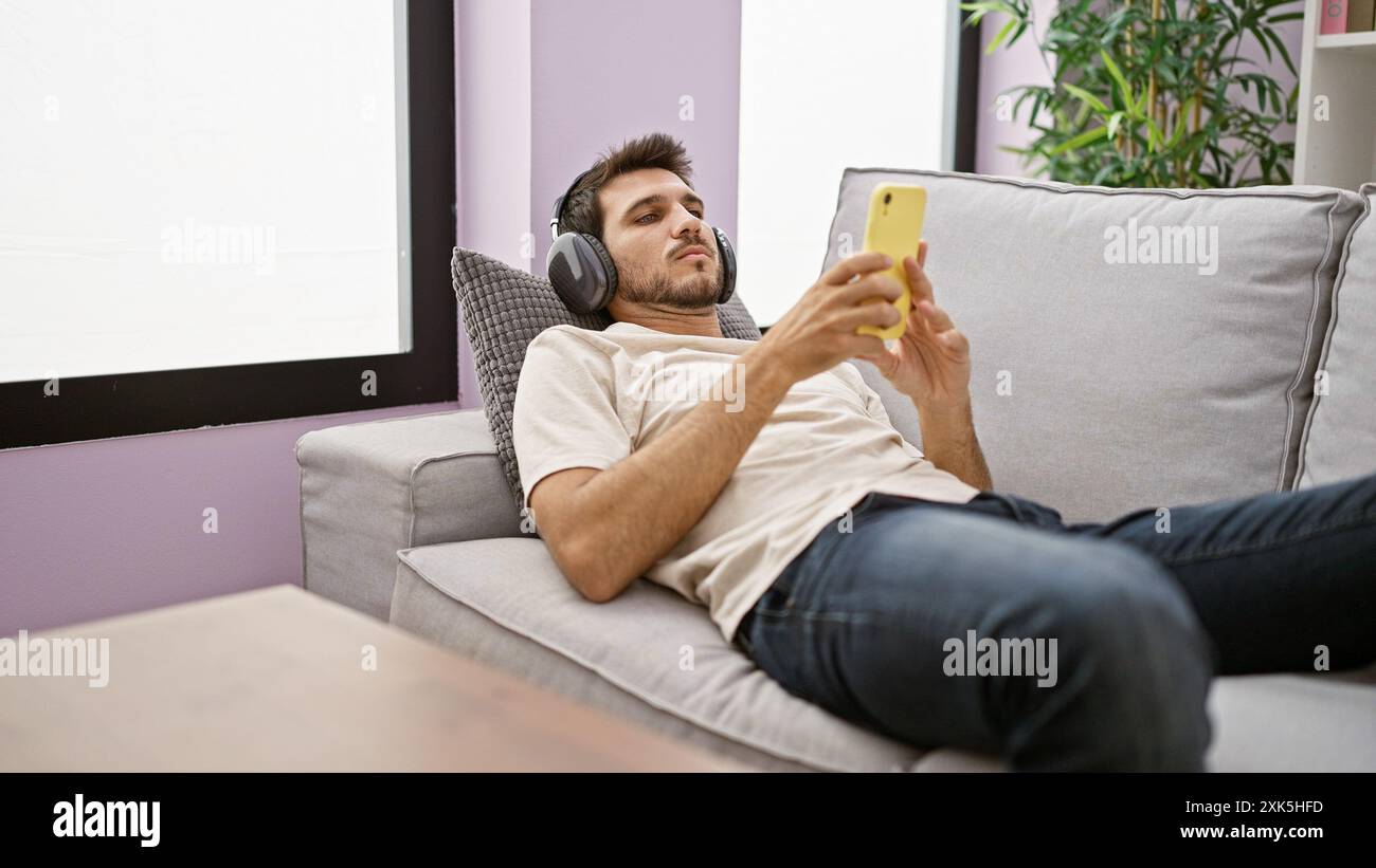 Un giovane ispanico con la barba si rilassa a casa, sdraiato sul divano mentre utilizza uno smartphone e indossa le cuffie. Foto Stock