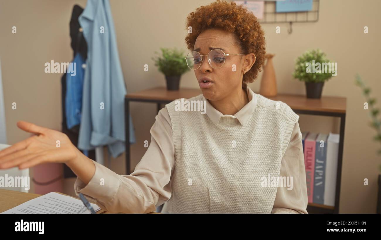 Donna afroamericana in un maglione che fa gesti mentre parla in casa con piante d'appartamento e scaffali in sottofondo. Foto Stock