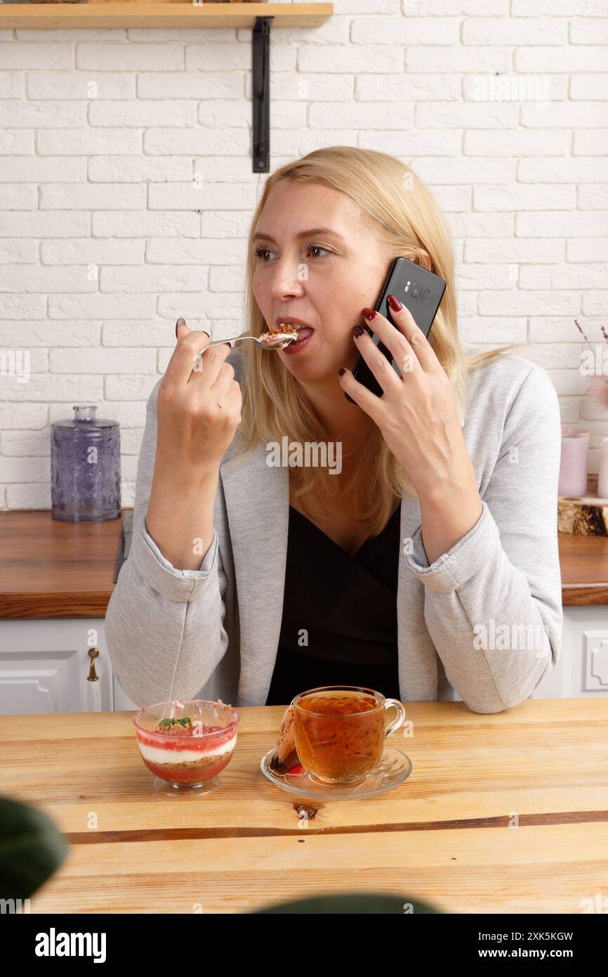 Donna bionda in giacca grigia al tavolo della cucina che mangia dessert, parla al telefono. Stile di vita moderno, alimentazione sana, relax, elegante, riunione di lavoro Foto Stock