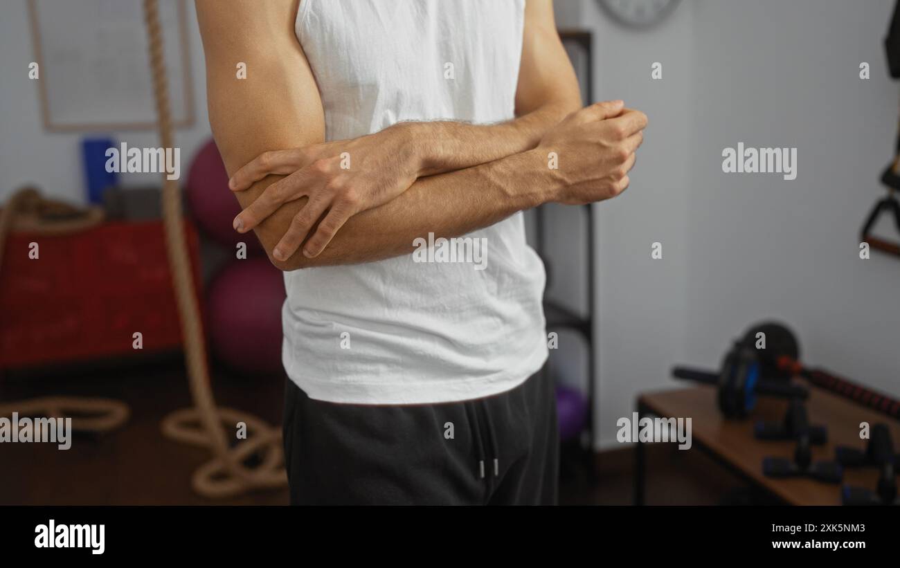 Uomo ispanico in piedi al chiuso in una palestra con le braccia incrociate indossando una canotta bianca e circondato da attrezzature e corde per l'esercizio fisico Foto Stock