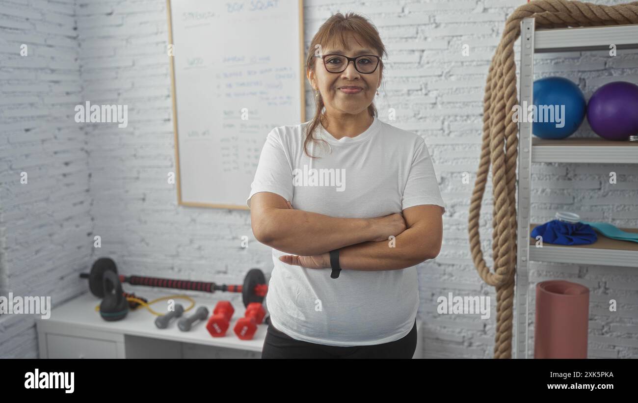 Una donna ispanica matura si trova in una palestra coperta con pareti bianche, braccia incrociate, circondata da attrezzature per il fitness come manubri e corde. Foto Stock