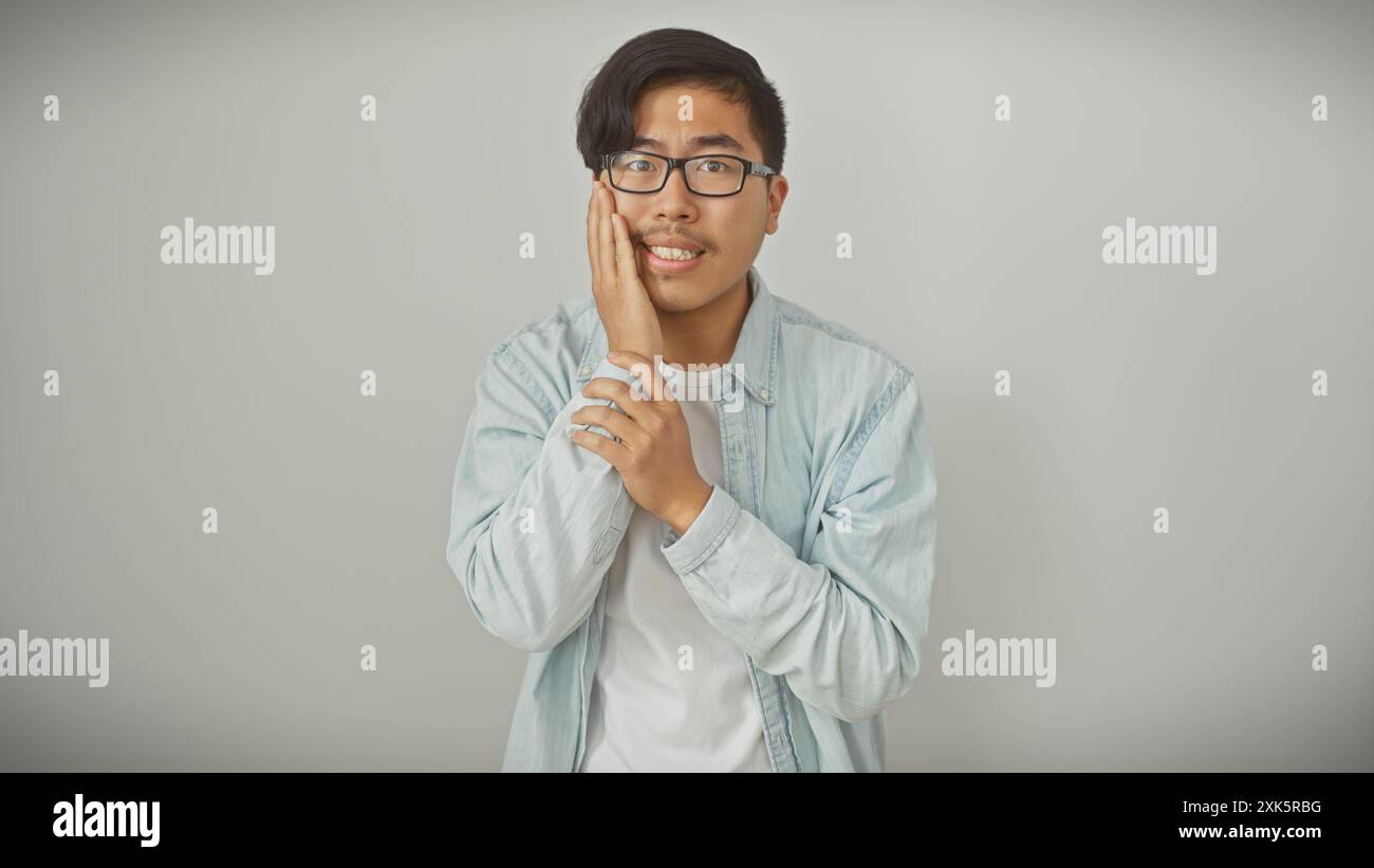 Un giovane uomo asiatico con occhiali in posa davanti a uno sfondo bianco, vestito casualmente con una giacca leggera in denim. Foto Stock