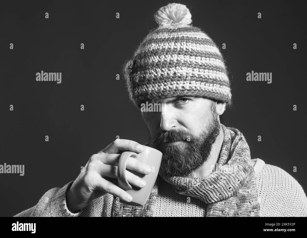 Un uomo serio ed elegante in maglione e cappello avvolto in sciarpa con tazza di caffè o tè. Autunno o inverno. Ritratto di un uomo barbuto alla moda Foto Stock