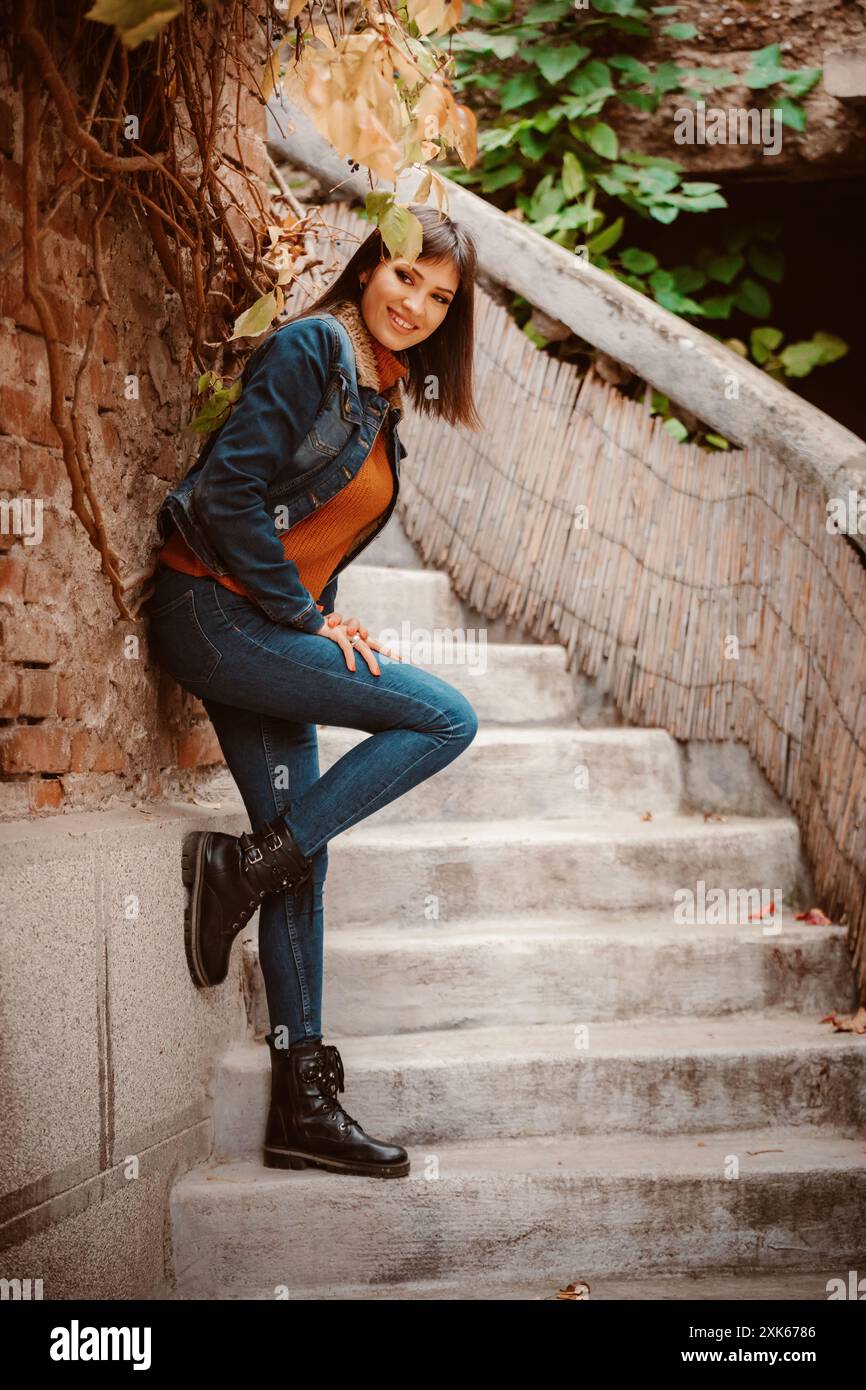Una giovane donna si appoggia a una parete di mattoni, indossando jeans blu e una giacca in denim mentre guarda la fotocamera. È in piedi su una serie di ste in cemento Foto Stock