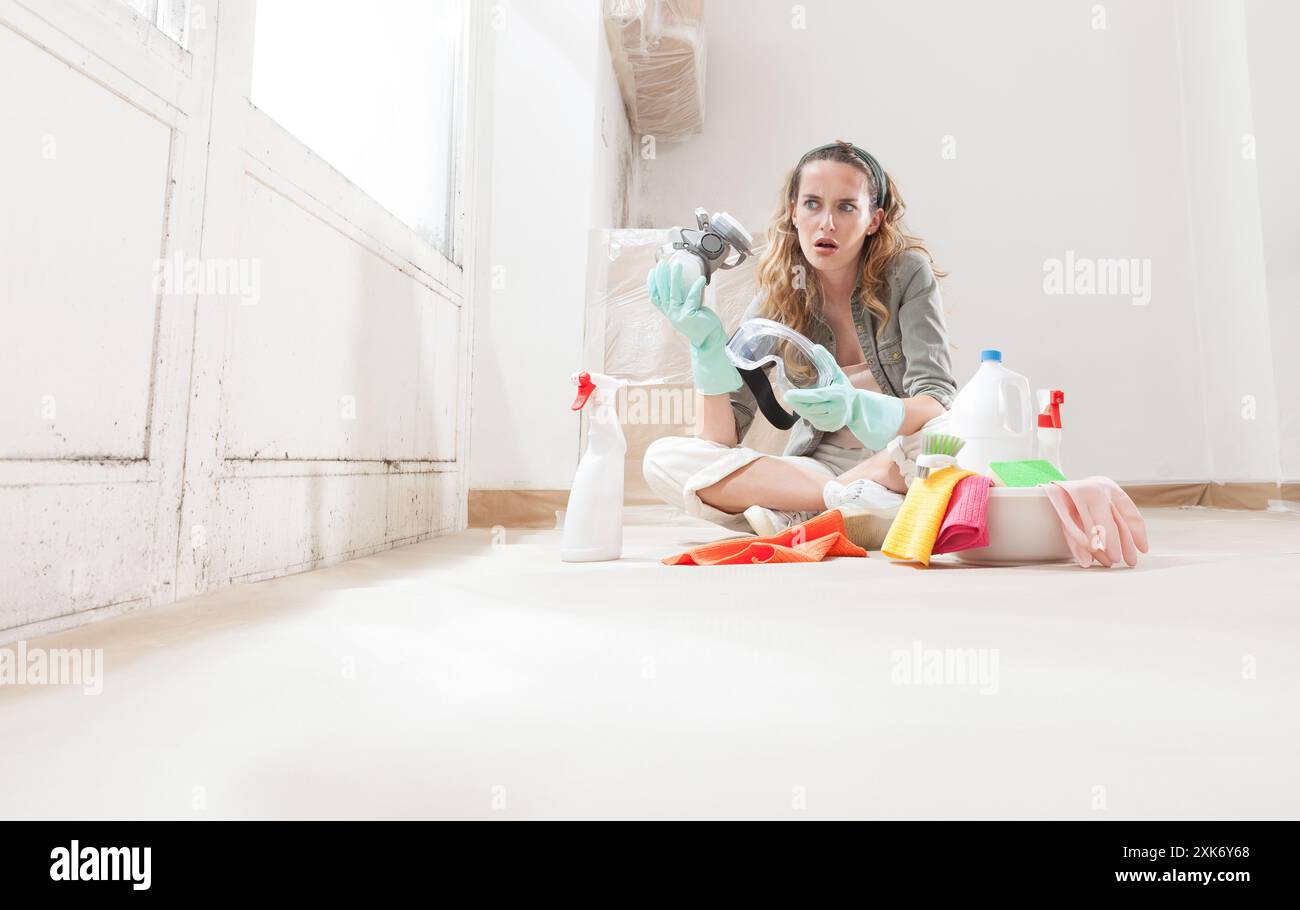 Una donna stanca e disgustata che pulisce la muffa sulla parete della finestra della casa regge una maschera respiratoria protettiva. Utilizzo di prodotti per la pulizia della casa per Foto Stock