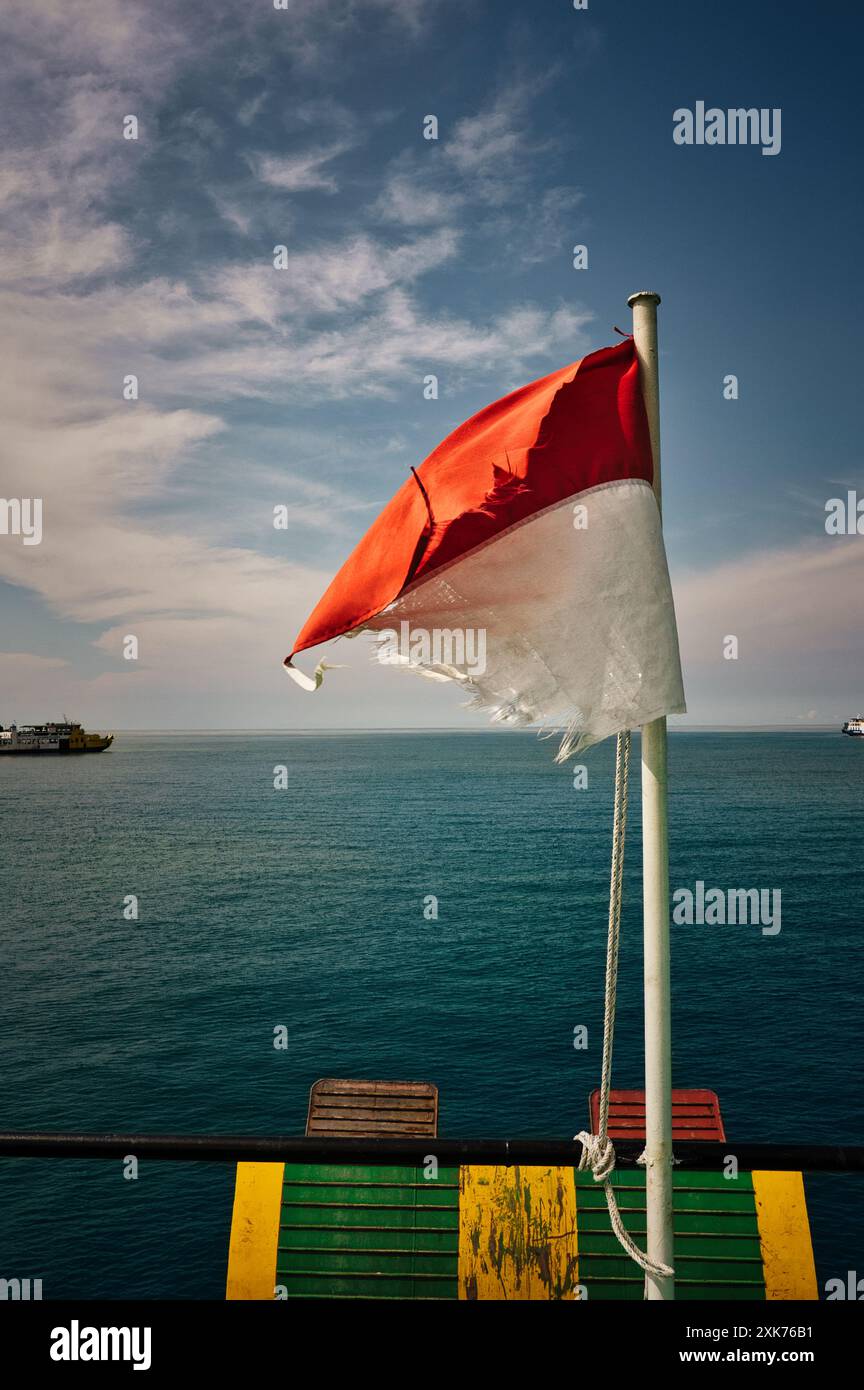 Bandiera indonesiana sul mare calmo, Una vecchia bandiera indonesiana che ondeggia nel vento su un tranquillo sfondo blu dell'oceano. Le barche galleggiano all'orizzonte. Foto Stock