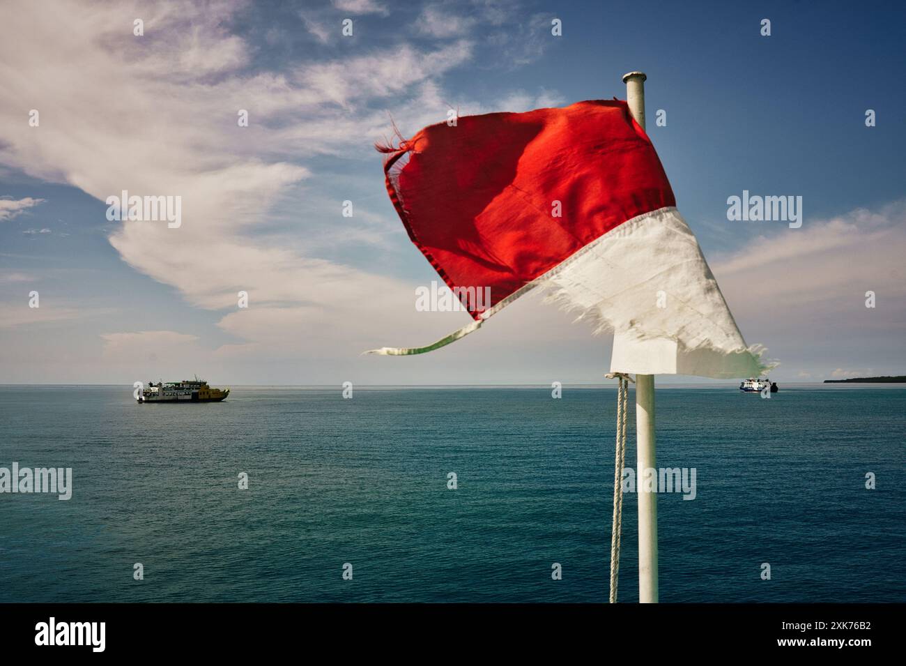 Bandiera indonesiana sul mare calmo, Una vecchia bandiera indonesiana che ondeggia nel vento su un tranquillo sfondo blu dell'oceano. Le barche galleggiano all'orizzonte. Foto Stock