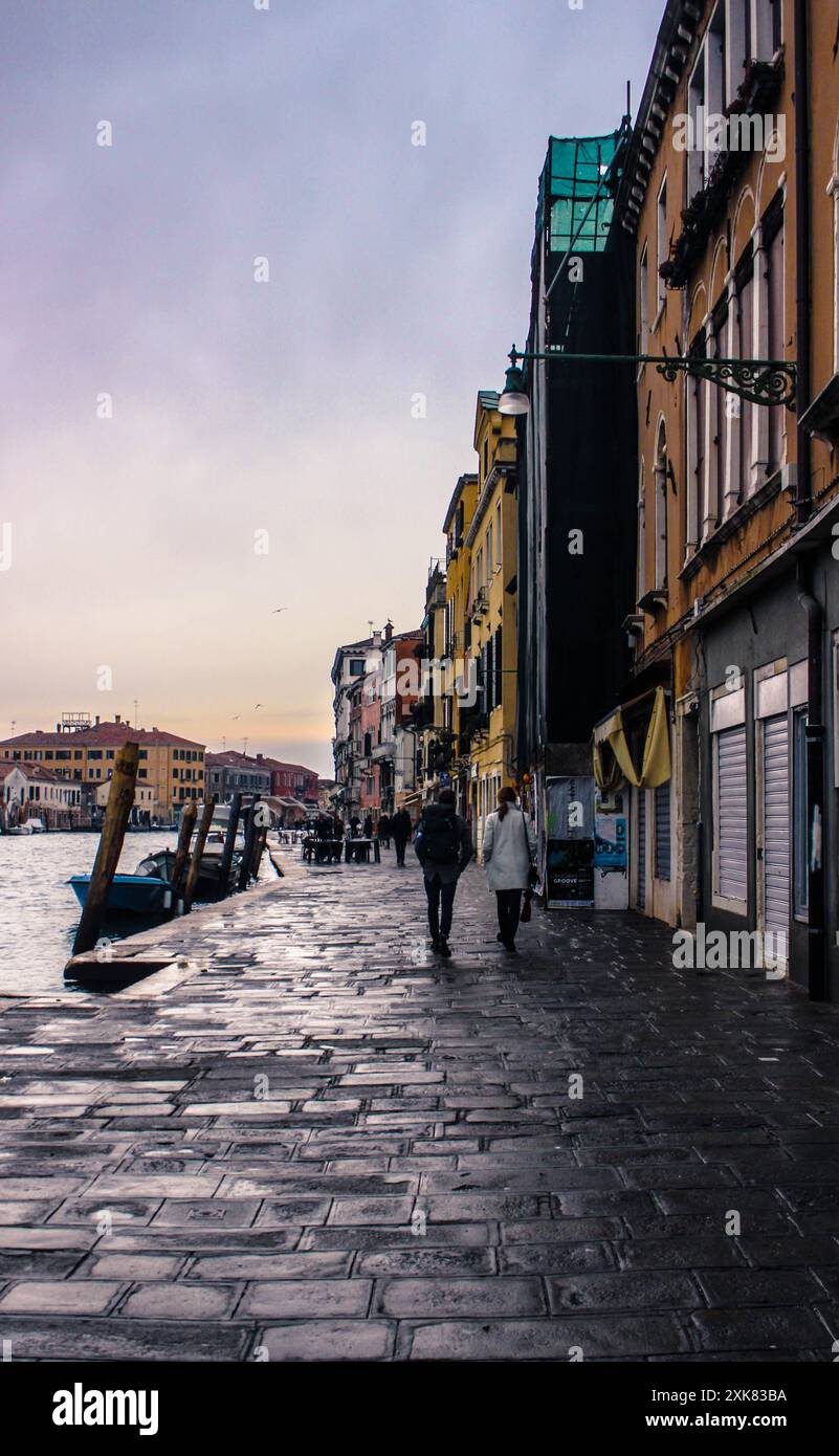 Due persone camminano lungo le strade acciottolate di Venezia, Italia, con canali e architettura storica sullo sfondo. L'atmosfera è tranquilla Foto Stock