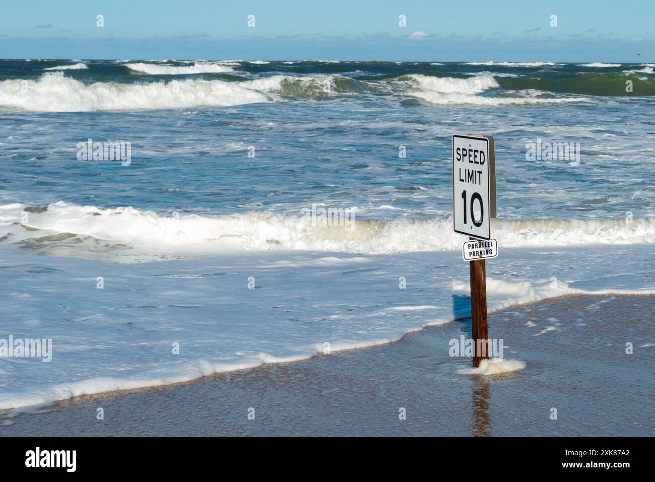 C'è un limite di velocità di 10 segnali e un segnale di parcheggio parallelo su un palo di legno su una spiaggia di sabbia. C'è una spiaggia tempestosa con onde e correnti che si muovono sulla riva. Foto Stock