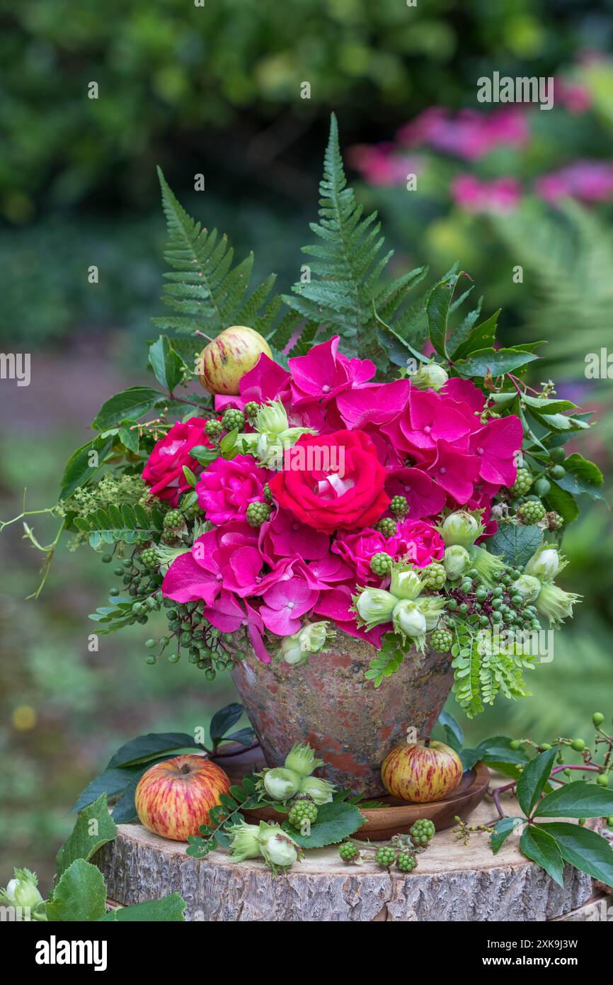 composizione floreale con rose rosa, ortensie in fiore, nocciole, more e foglie in una pentola di terracotta d'epoca in giardino Foto Stock