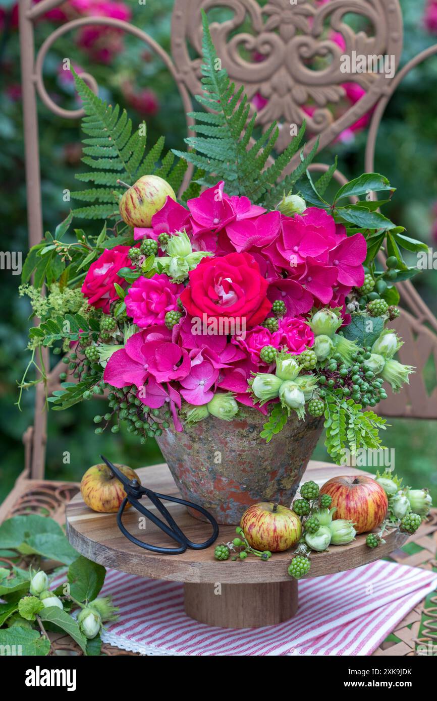 composizione floreale con rose rosa, ortensie in fiore, nocciole, more e foglie in una pentola di terracotta d'epoca in giardino Foto Stock