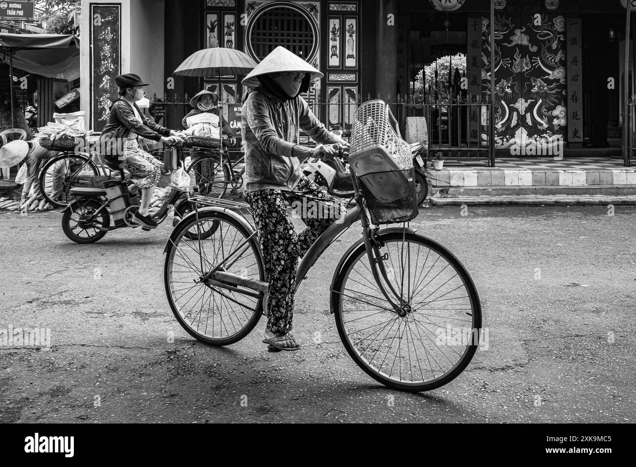 Una donna anziana con un cappello conico in bicicletta a Hoi An Vietnam. Donna vietnamita in tradizionale cappello di bambù in bicicletta lungo l'antica città di Hoi An. Trav Foto Stock