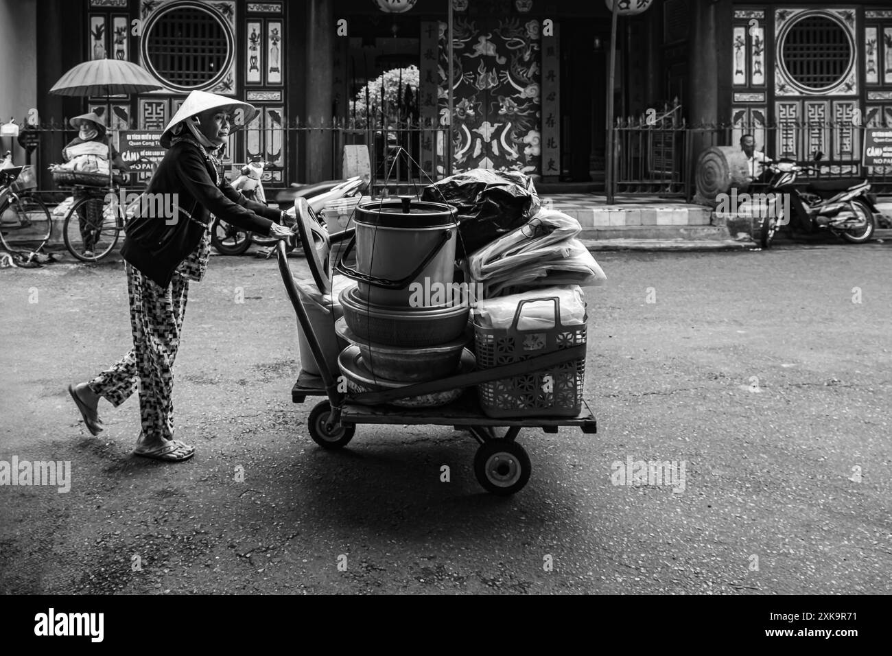 Vecchia vietnamita che spinge un carro con le merci. Hoi An, Vietnam. Una donna che indossa abiti tradizionali vietnamiti spingendo un trolley in metallo. Foto di viaggio i Foto Stock