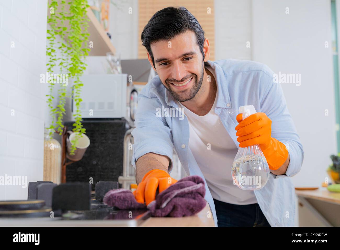 Pulisci le tue preoccupazioni. un giovane che disinfetta un bancone da cucina a casa. Giovane e affascinante uomo con grembiule e guanti protettivi, lavello e rubinetto Foto Stock