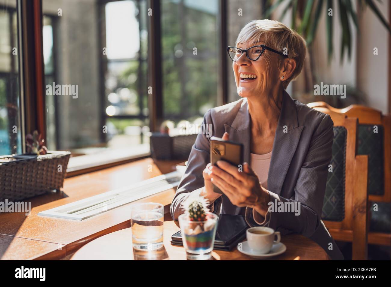 Una donna matura è seduta in una caffetteria e si sta rilassando. Sta bevendo caffè e sta messaggiando al telefono. Foto Stock