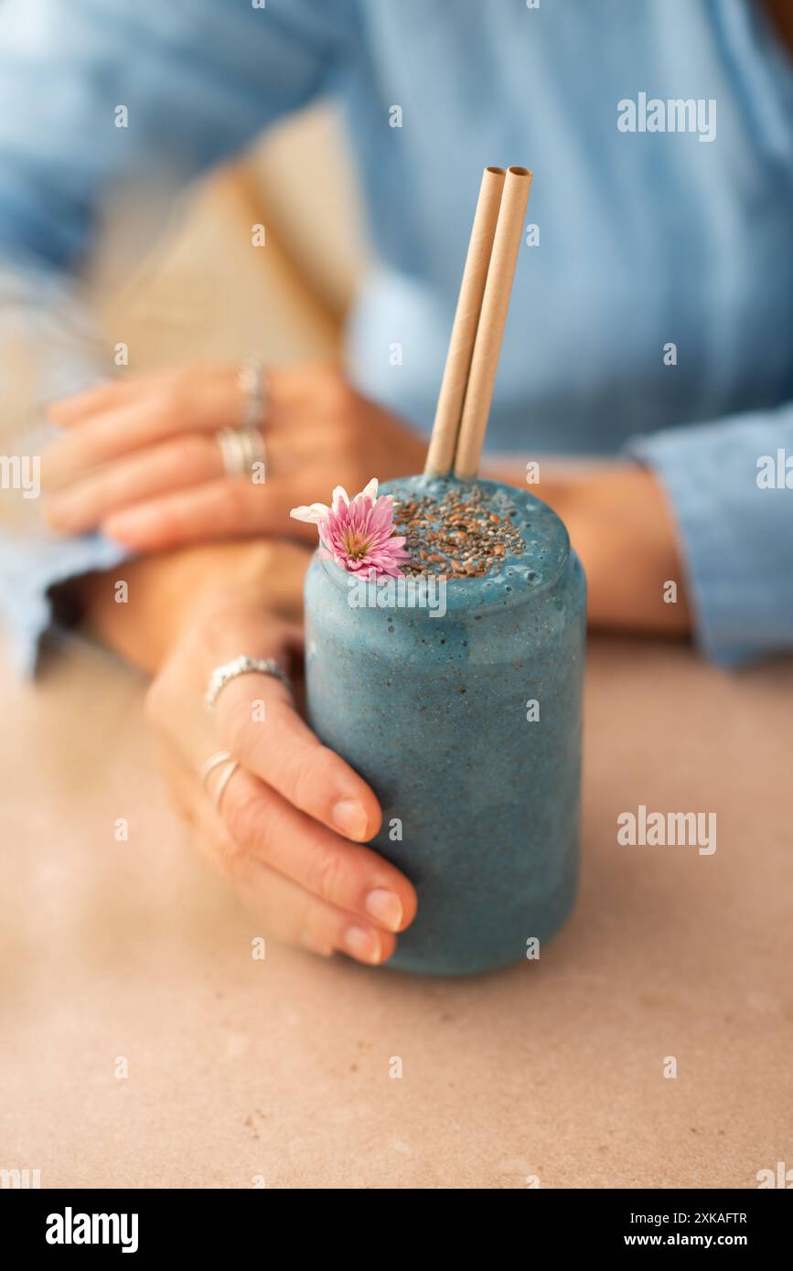 Frullato vegano spirulina blu con semi di chia, decorato con fiori commestibili. Donna che regge un bicchiere di frullato vegano. Foto Stock