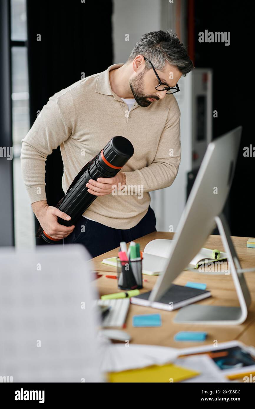 Un uomo d'affari con la barba che indossa gli occhiali si trova in un ufficio moderno con un grande schermo per computer, guardando un tubo che tiene in mano. Foto Stock