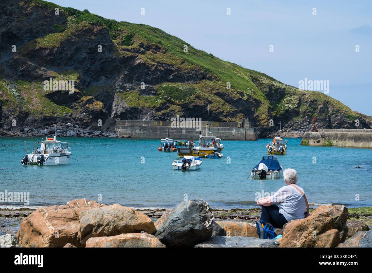 Signora matura seduta su una roccia sulla spiaggia del porto osservando un pescatore che lavora sulla sua barca in una giornata estiva di sole Port Isaac, Cornovaglia, Inghilterra, Regno Unito Foto Stock