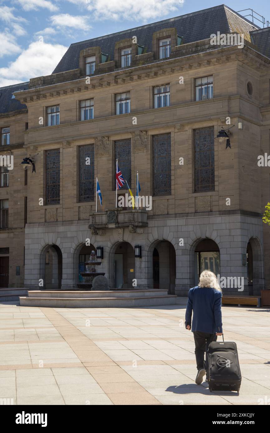 La persona (capelli lunghi e leggeri, che indossa un abito) che tira un trolley/valigia si dirige verso le Dundee City Chambers, sede del Consiglio comunale di Dundee Foto Stock