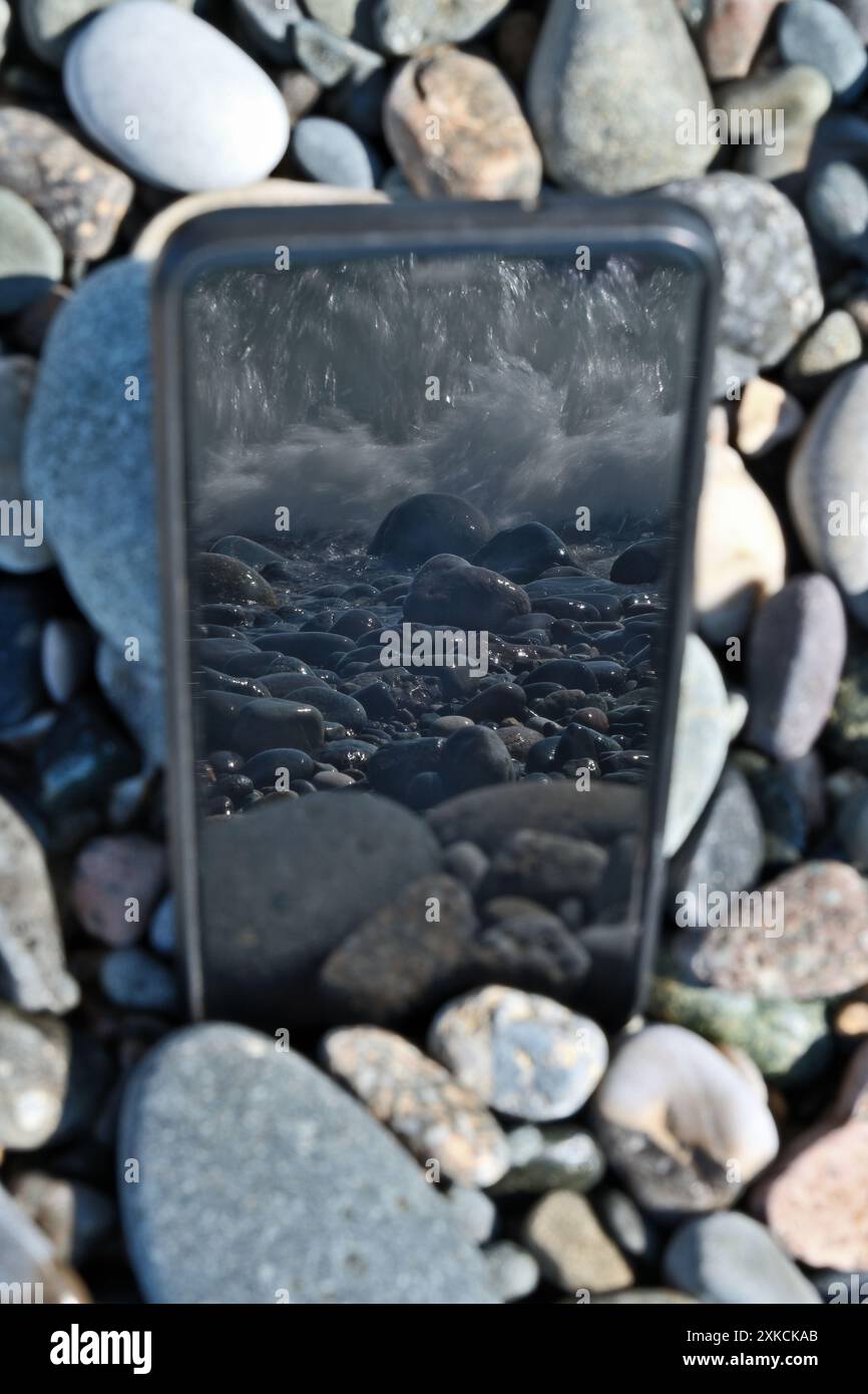 Telefono cellulare con un riflesso del mare su una spiaggia di ciottoli Foto Stock