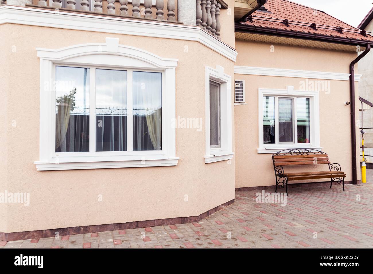 La facciata di una casa presenta grandi finestre con cornici bianche, pareti beige e un tetto piastrellato di rosso. Una panca in legno con dettagli in ferro battuto è plac Foto Stock