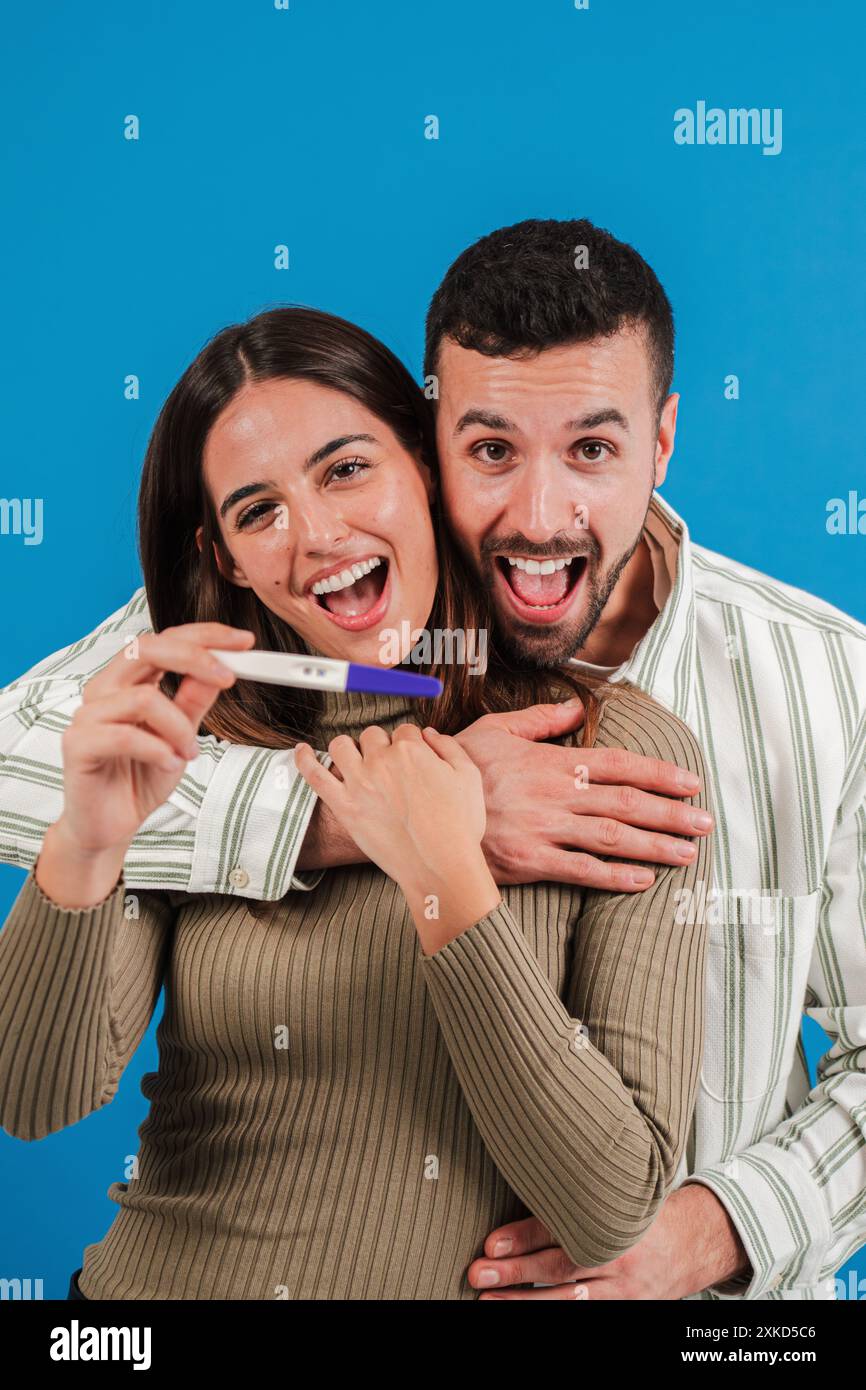 Verticale. Una felice coppia caucasica che mostra un test di gravidanza positivo alla telecamera, celebrando che saranno genitori su uno sfondo blu. Genitorialità, maternità e paternità. Foto di alta qualità Foto Stock