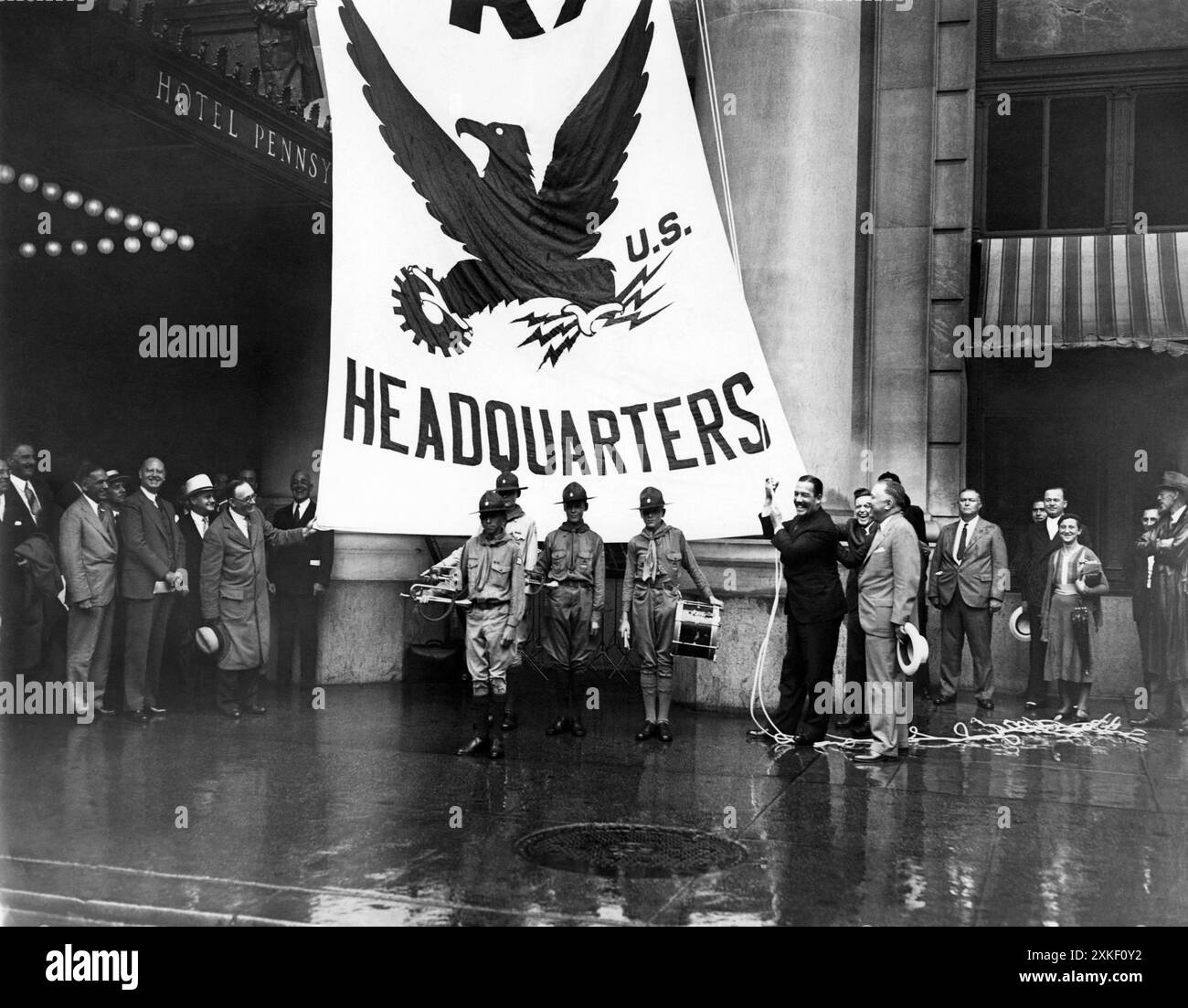 New York, New York 21 agosto 1933 nemmeno un downpour poteva fermare le cerimonie di innalzamento della bandiera dell'NRA presso l'Hotel Pennsylvania, dato che un enorme striscione della National Recovery Administration è stato innalzato in una posizione di rilievo davanti all'hotel. Grover Whalen, presidente della campagna di New York City, è mostrato mentre officiava, mentre diversi funzionari della città e una piccola folla, tenuti al minimo dalla pioggia, guardavano. Foto Stock