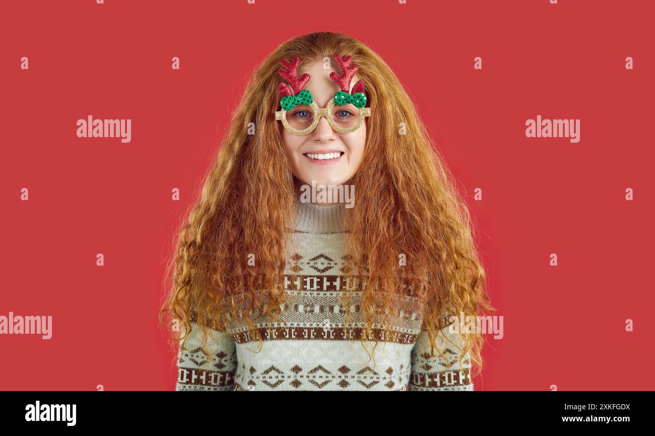 Giocosa ragazza rossa felice con maglione natalizio e occhiali con corna di cervo su sfondo rosso. Foto Stock