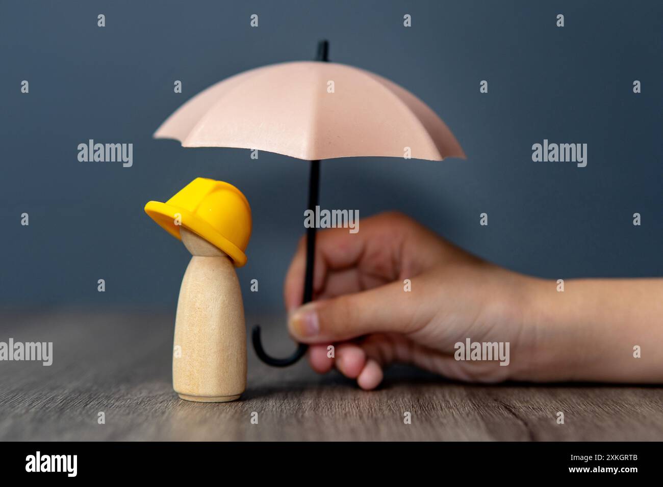 Immagine ravvicinata della mano che regge un ombrello su una bambola di legno con un elmetto giallo. Concetto di assicurazione per lavoratori e dipendenti. Foto Stock