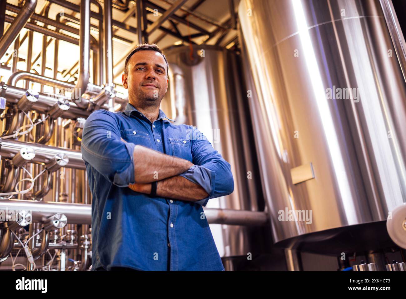 Un giovane uomo sorridente in abiti casual è in piedi nell'officina della fabbrica. Un bel lavoratore lavora in uno stabilimento. Un uomo in una birreria. Produzione e Foto Stock