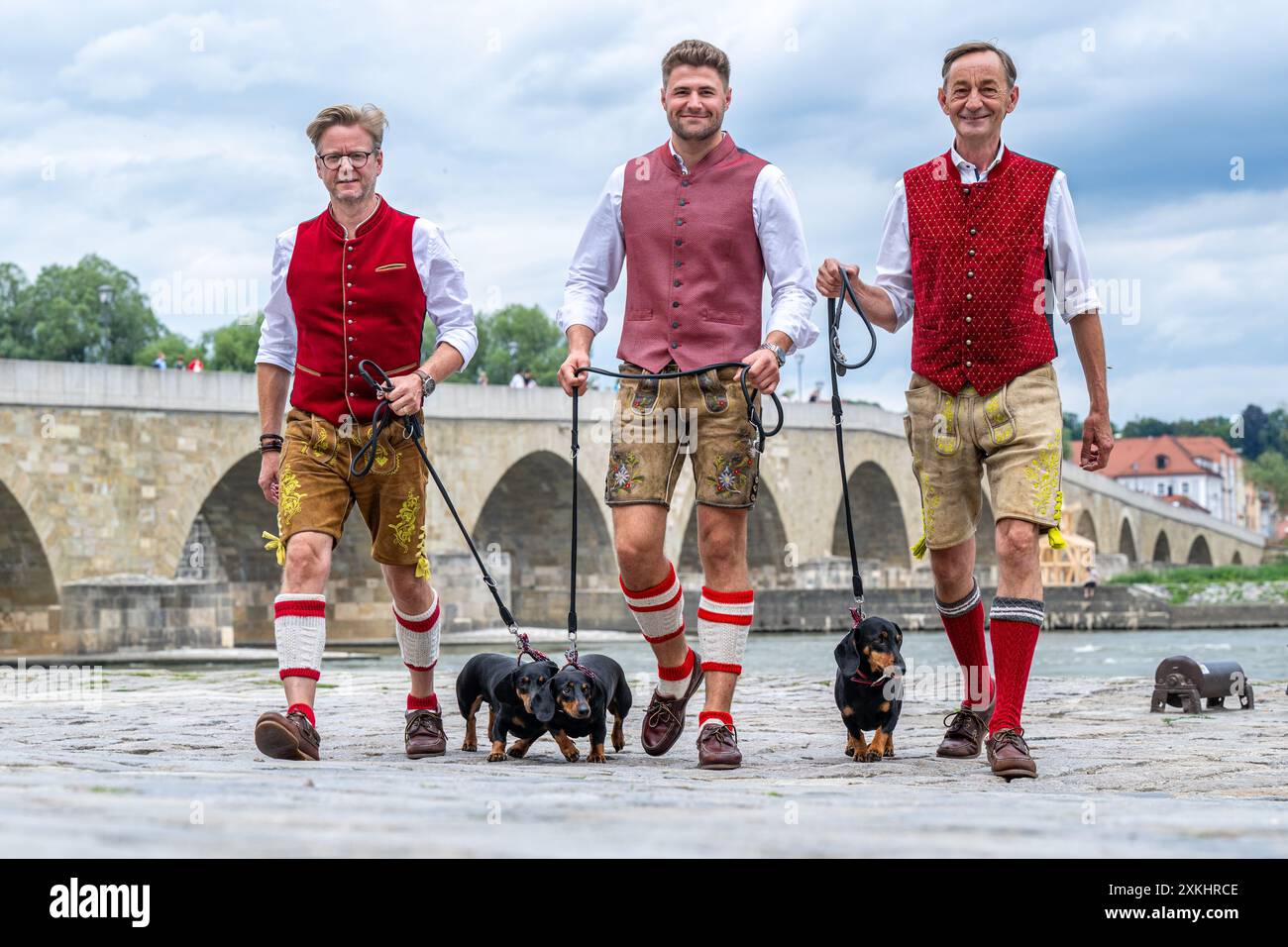 Ratisbona, Germania. 23 luglio 2024. Seppi Küblbeck (l-r) Moritz Hickl e Oliver Storz dal Museo Dachshund camminano con tre distese di fronte al Ponte di pietra sul Danubio. Crediti: Armin Weigel/dpa/Alamy Live News Foto Stock