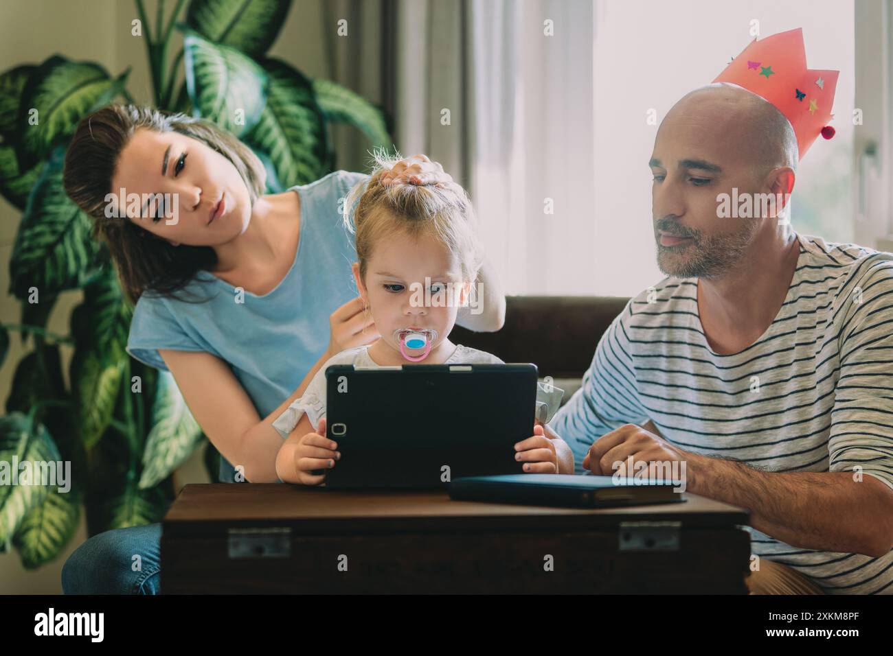 Una famiglia giovane è riunita intorno a un tablet digitale, per godersi del tempo insieme durante l'apprendimento e il gioco. I genitori sono impegnati ad aiutare i loro Foto Stock