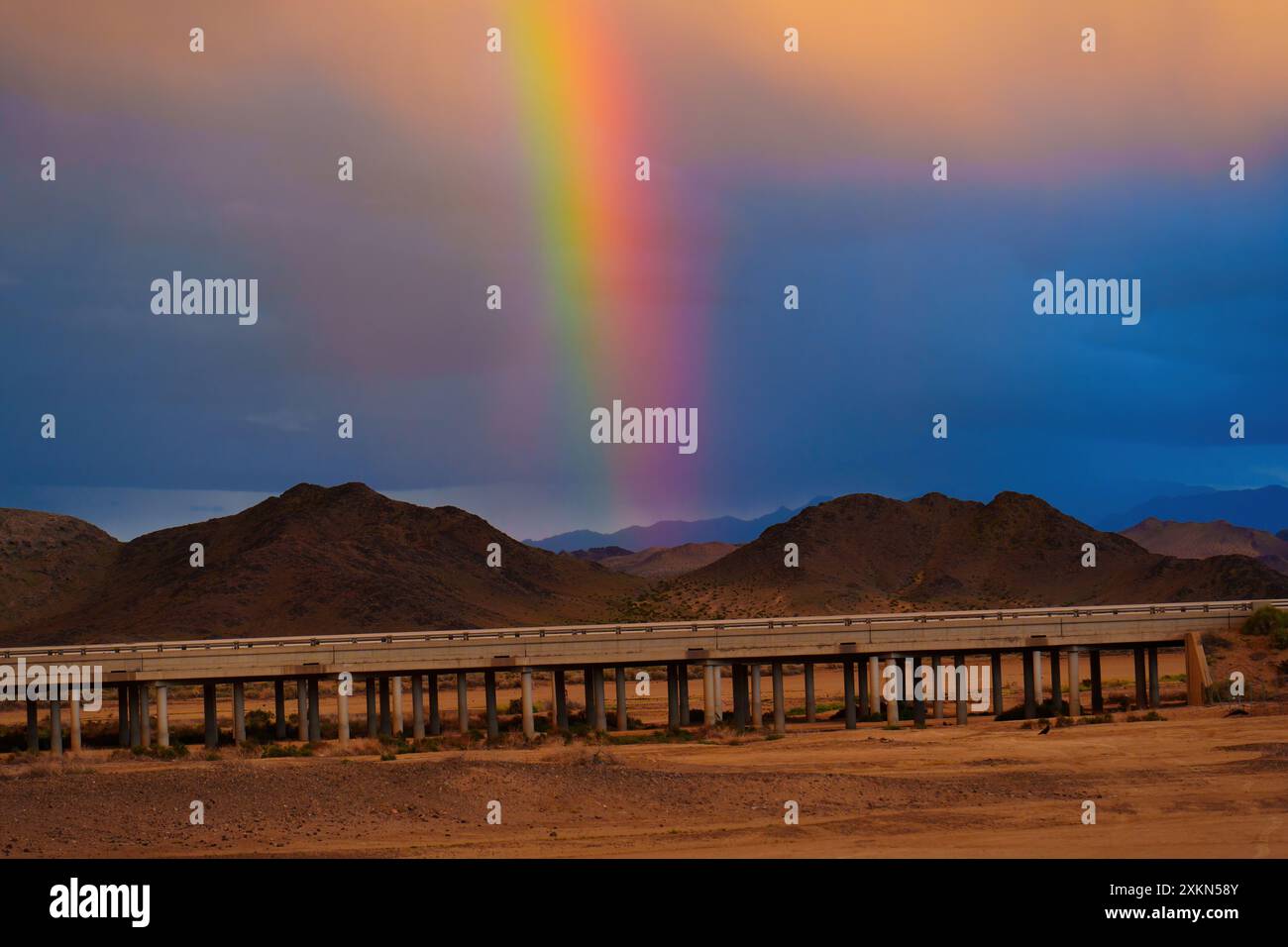 Il luminoso arcobaleno tocca l'arido deserto californiano, mostrando colori vivaci. La carreggiata sopraelevata si estende su terreni accidentati, sotto un cielo spettacolare Foto Stock