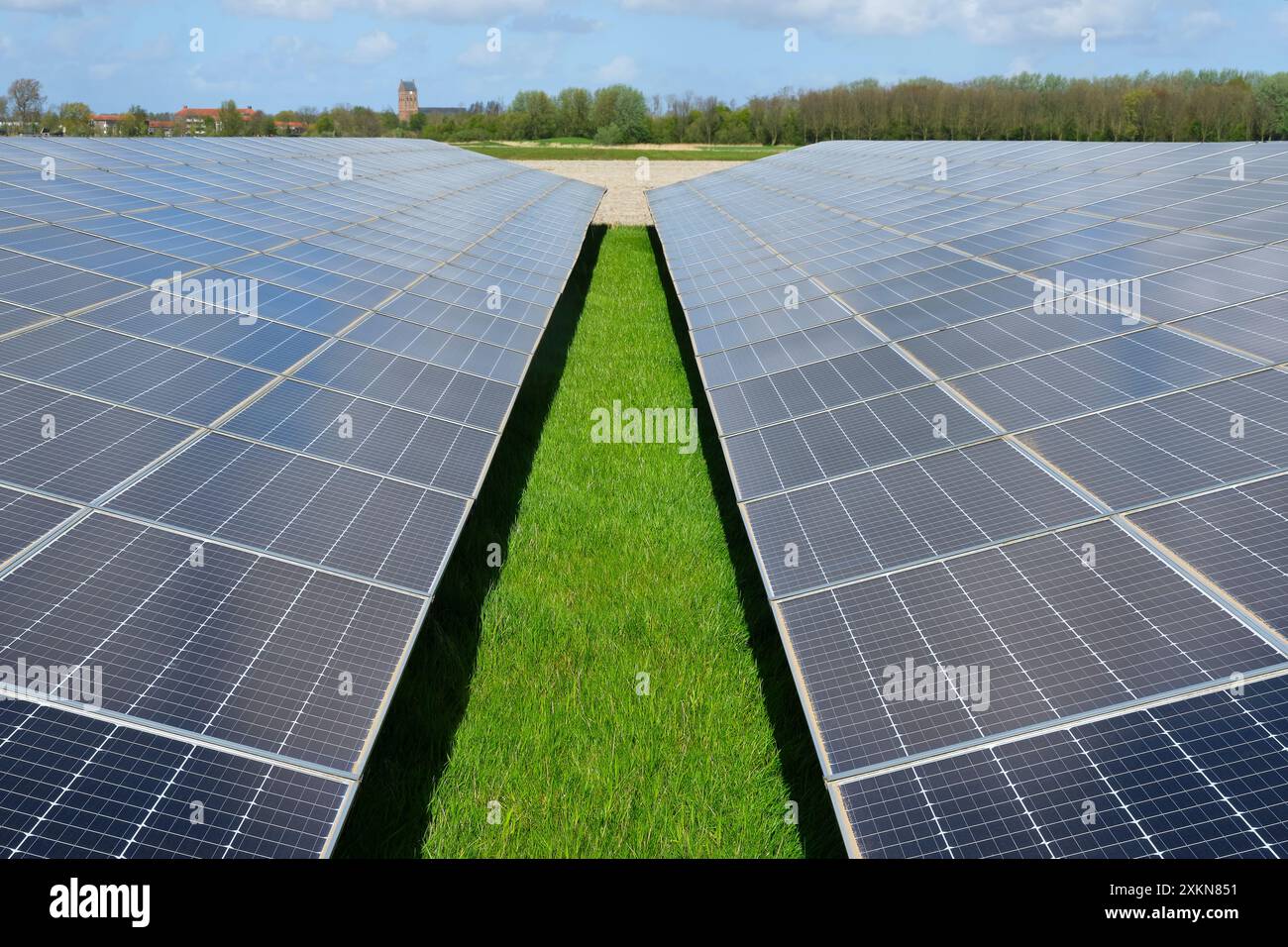 Celle solari blu in una centrale elettrica a pannelli solari o in un parco di celle fotovoltaiche che riflettono il cielo nuvoloso blu in una zona rurale e in un piccolo villaggio nella parte posteriore Foto Stock